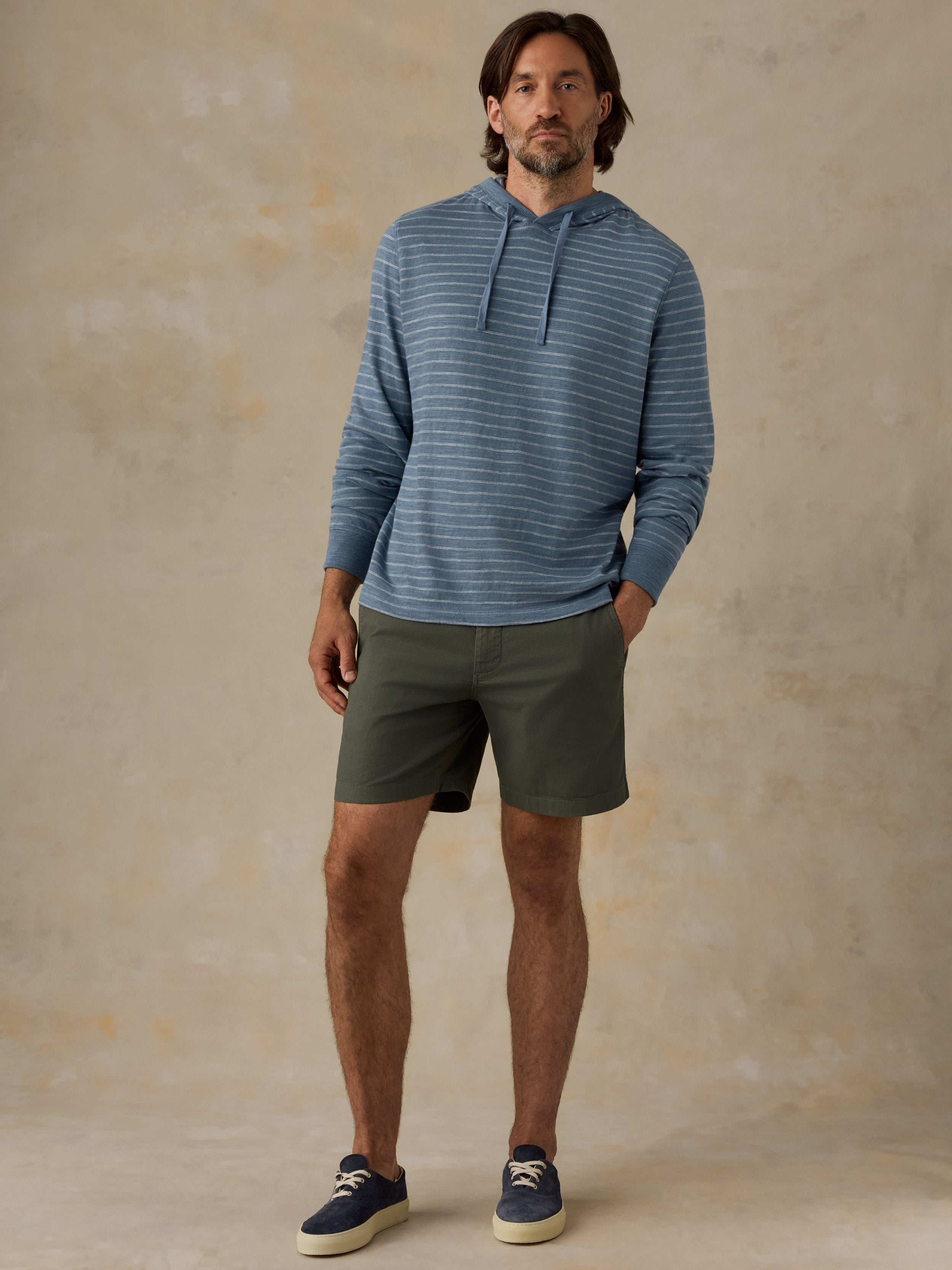 A man modeling a blue and white striped long-sleeve hoodie with olive green shorts and navy blue sneakers, standing against a textured beige studio backdrop.