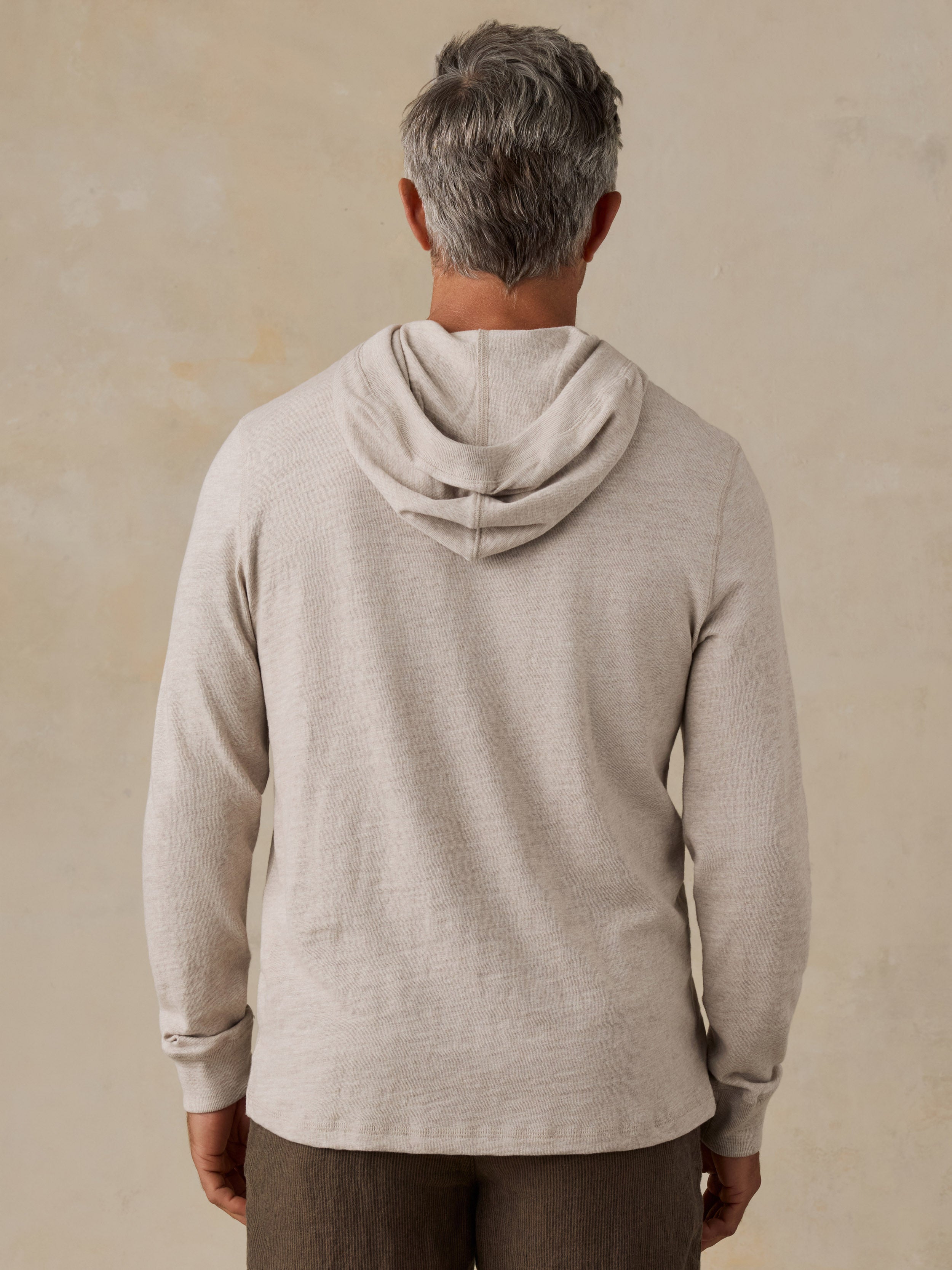 A man viewed from behind wearing a light beige heathered hooded long-sleeve shirt against a textured beige background.