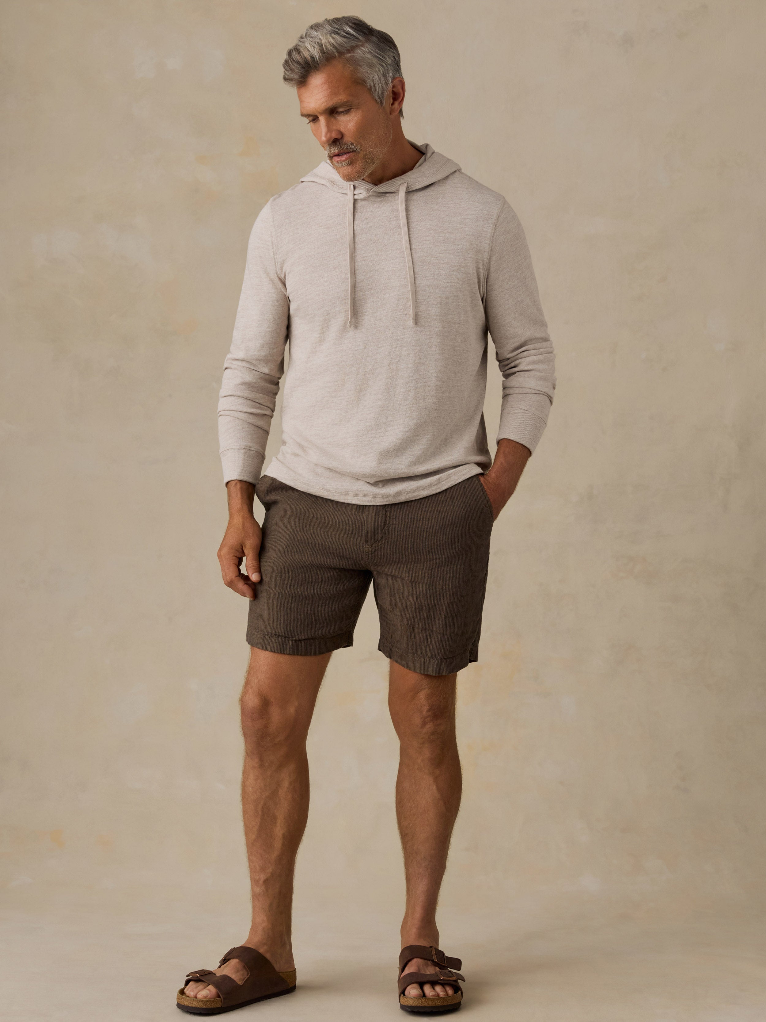 A man wearing a light beige long-sleeve hooded knit top, brown linen shorts, and brown sandals, standing against a textured beige studio backdrop.