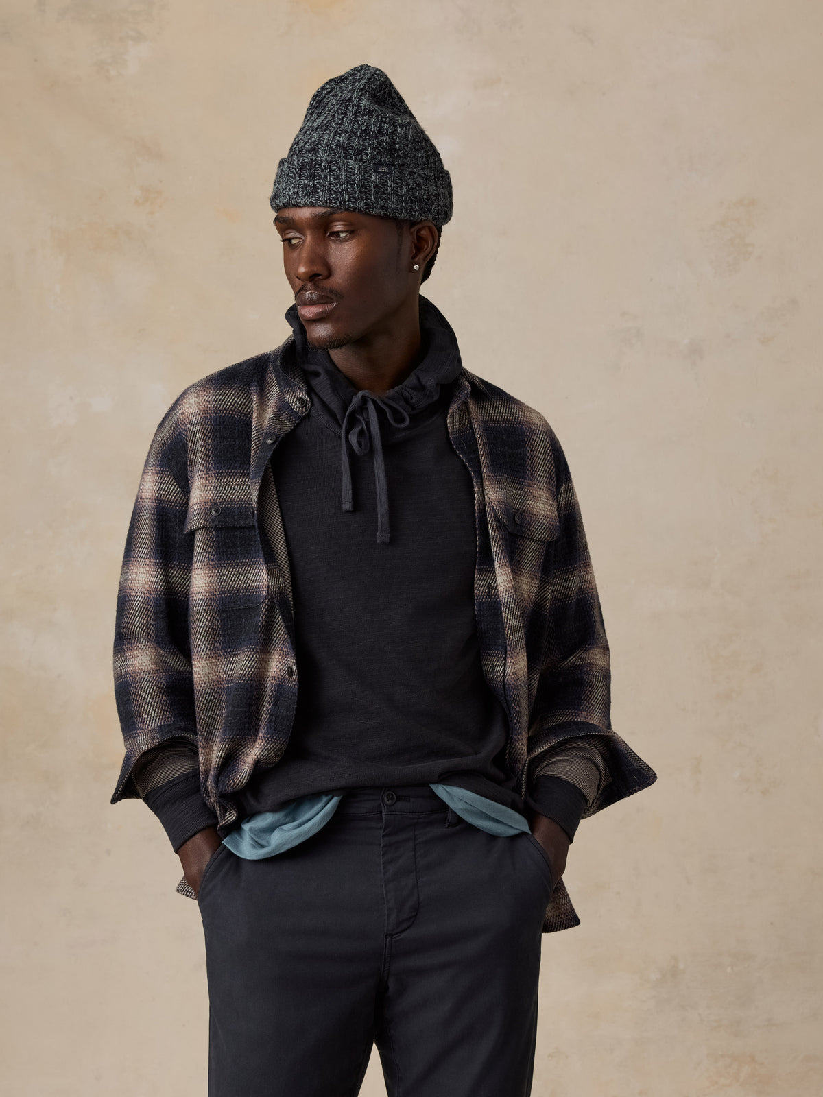 Man wearing a flannel shirt over a black hoodie and navy chino pants against a beige studio background, image 2 of 7