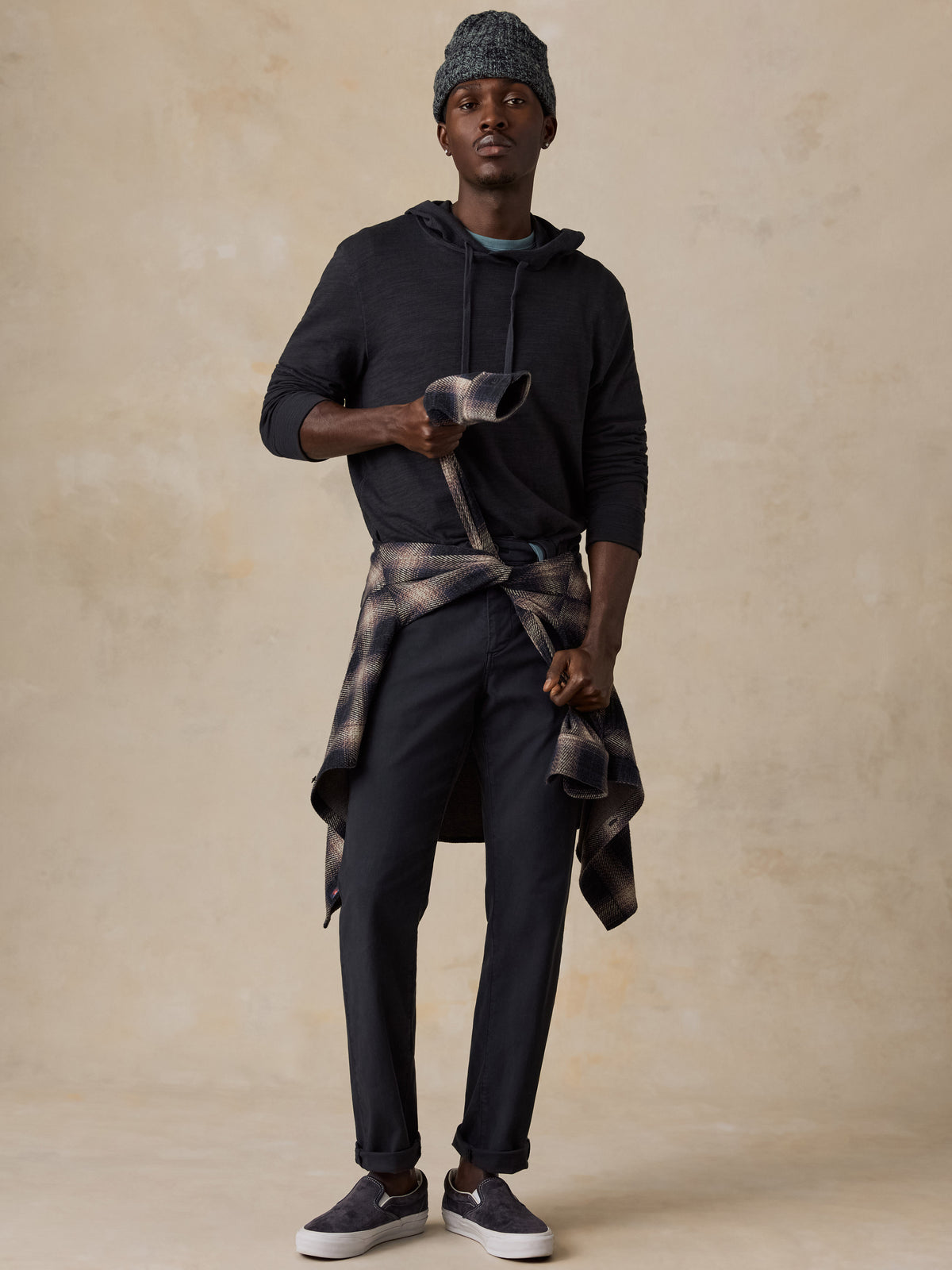 Man wearing a black hoodie over a blue t-shirt and navy chino pants with a flannel tied around his waist against a beige studio background, image 5 of 7