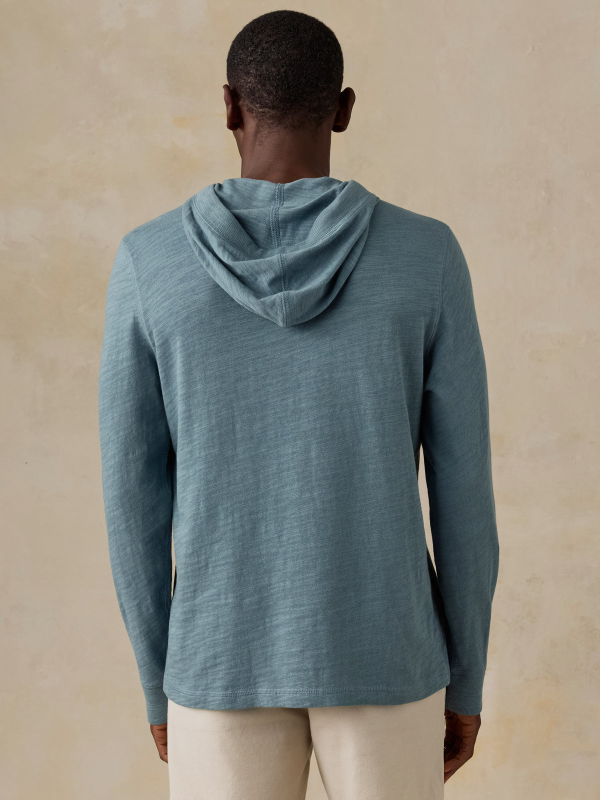 A man wearing a slate blue hooded long-sleeve shirt and cream pants.