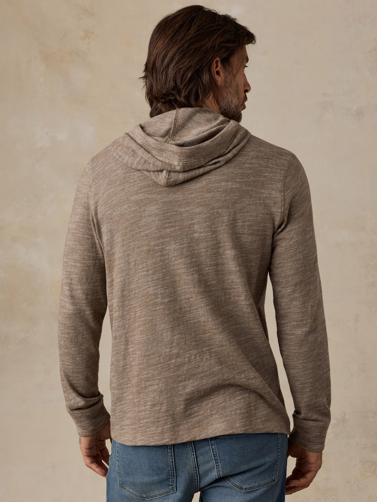A man viewed from behind wearing a heathered taupe-brown long-sleeve hoodie and blue jeans, image 3 of 5