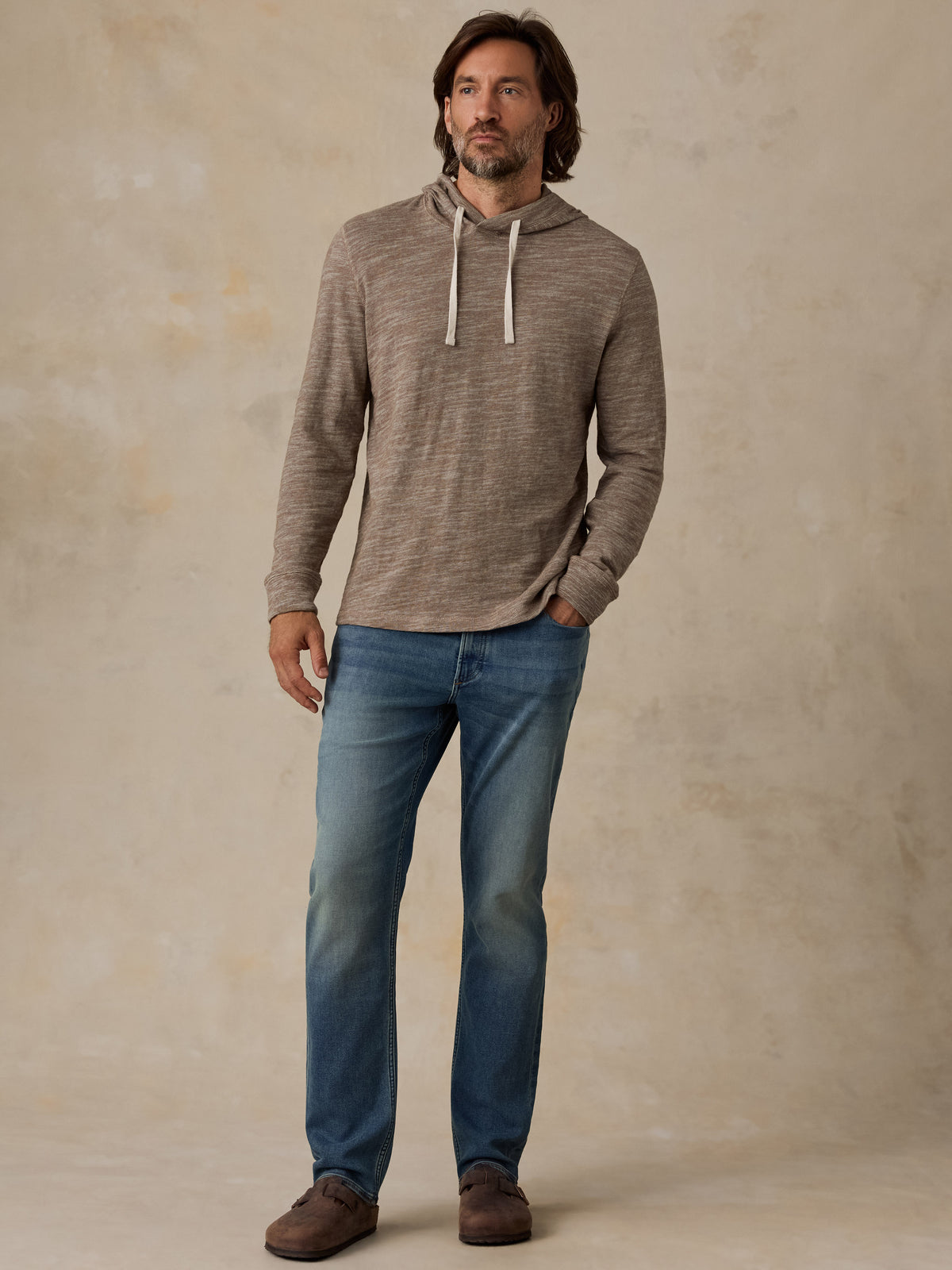 A man wearing a heathered taupe-brown long-sleeve hoodie and blue jeans, image 5 of 5
