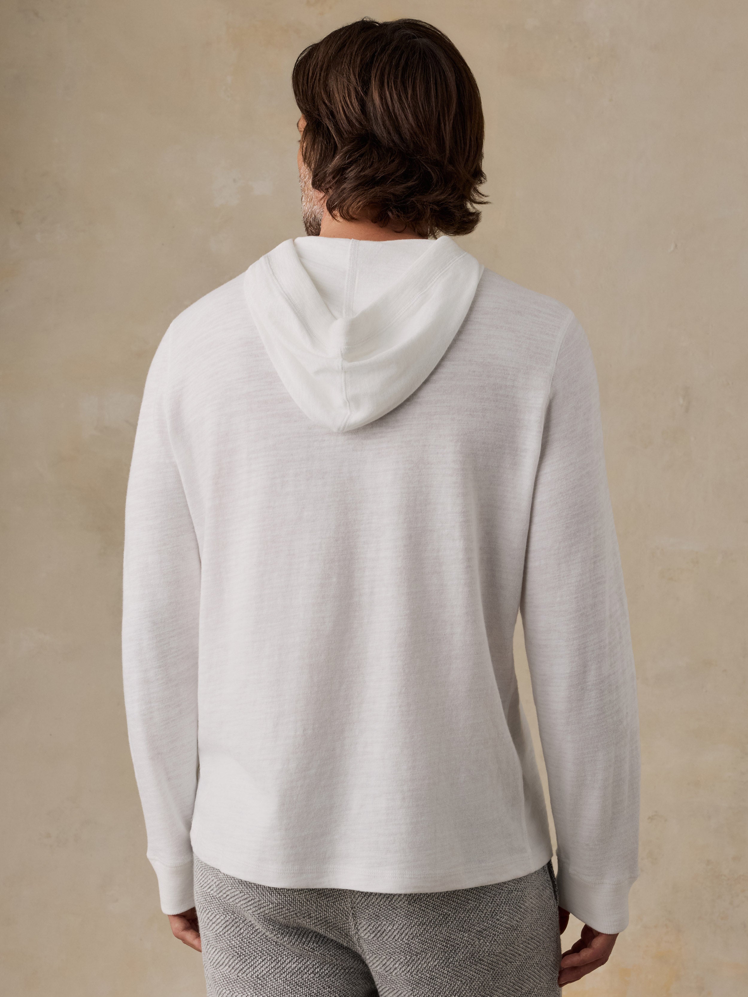 A man viewed from behind wearing a light off-white hoodie and gray textured pants, standing against a warm beige plaster wall.