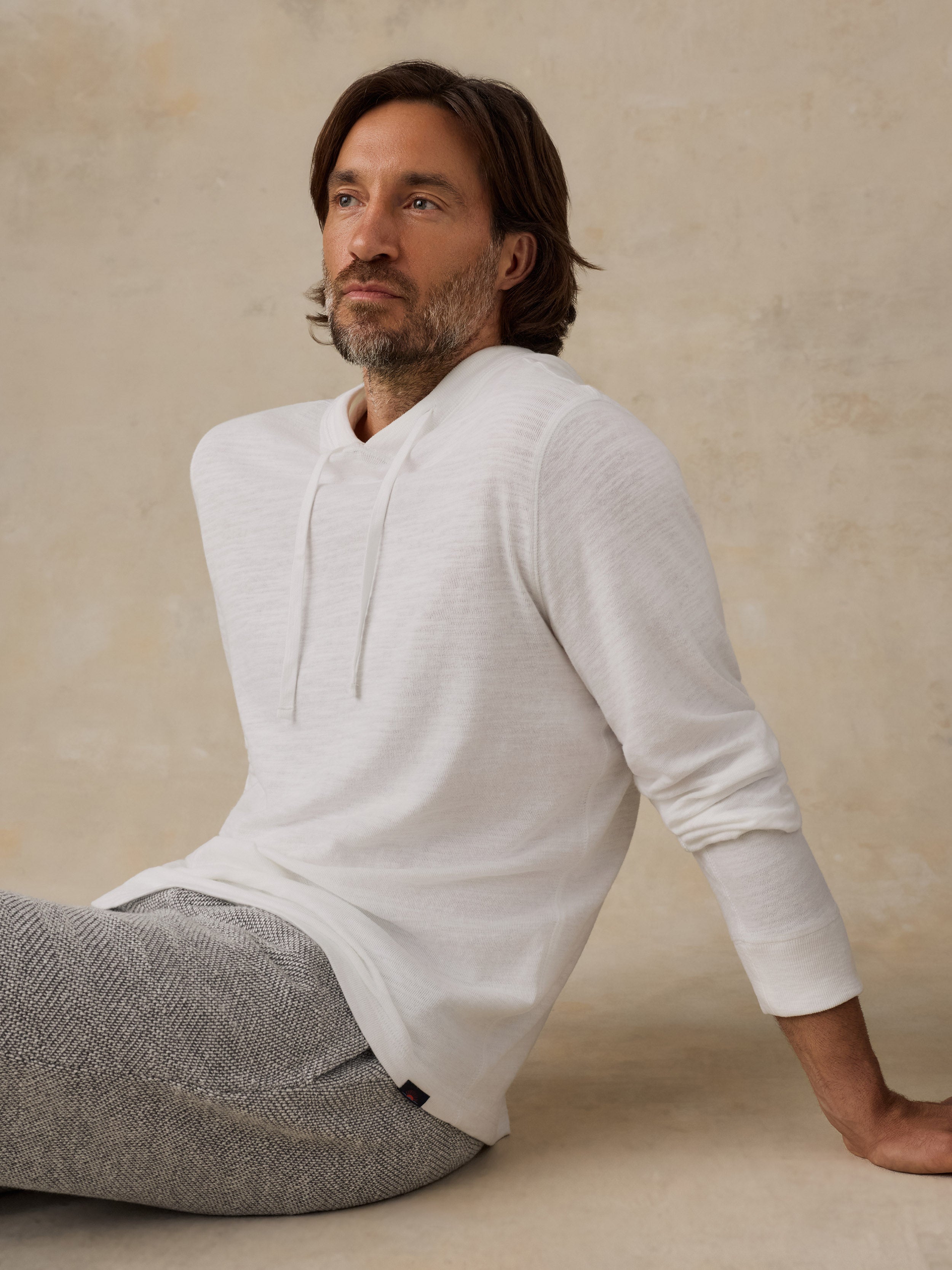A man wearing a white long-sleeve hoodie with a drawstring neckline and gray textured trousers, seated against a warm beige plaster wall.