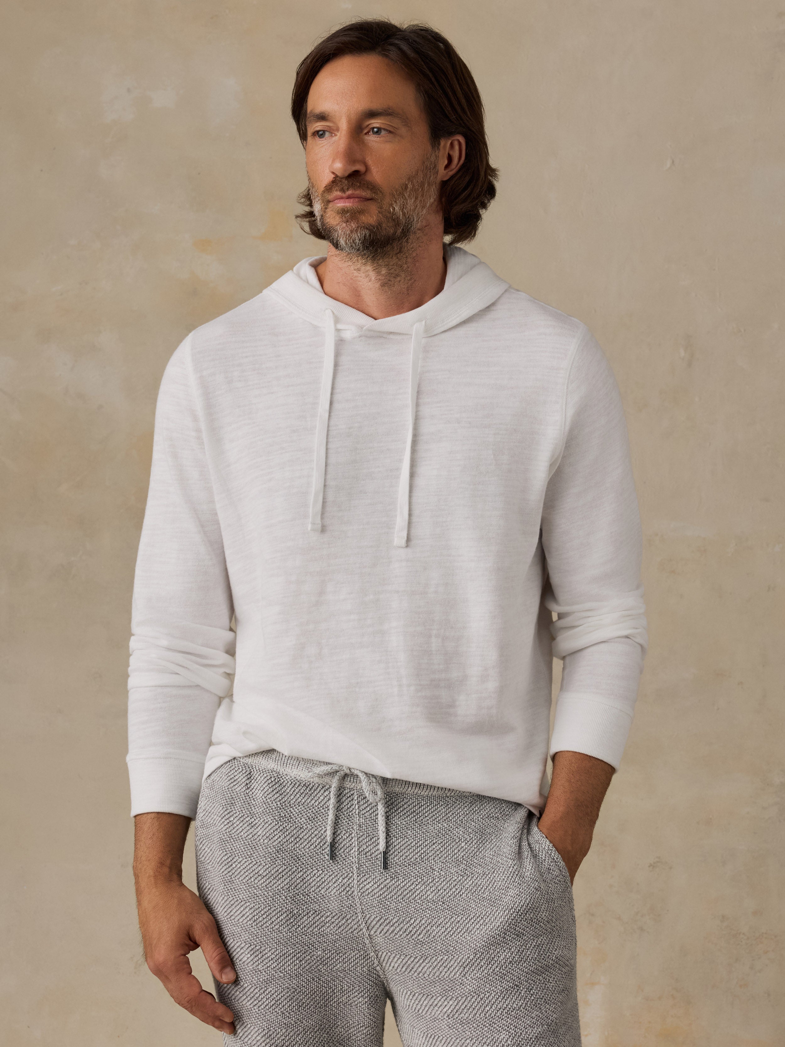 A man wearing a light off-white long-sleeve hoodie and heather gray drawstring sweatpants, standing against a textured beige wall.