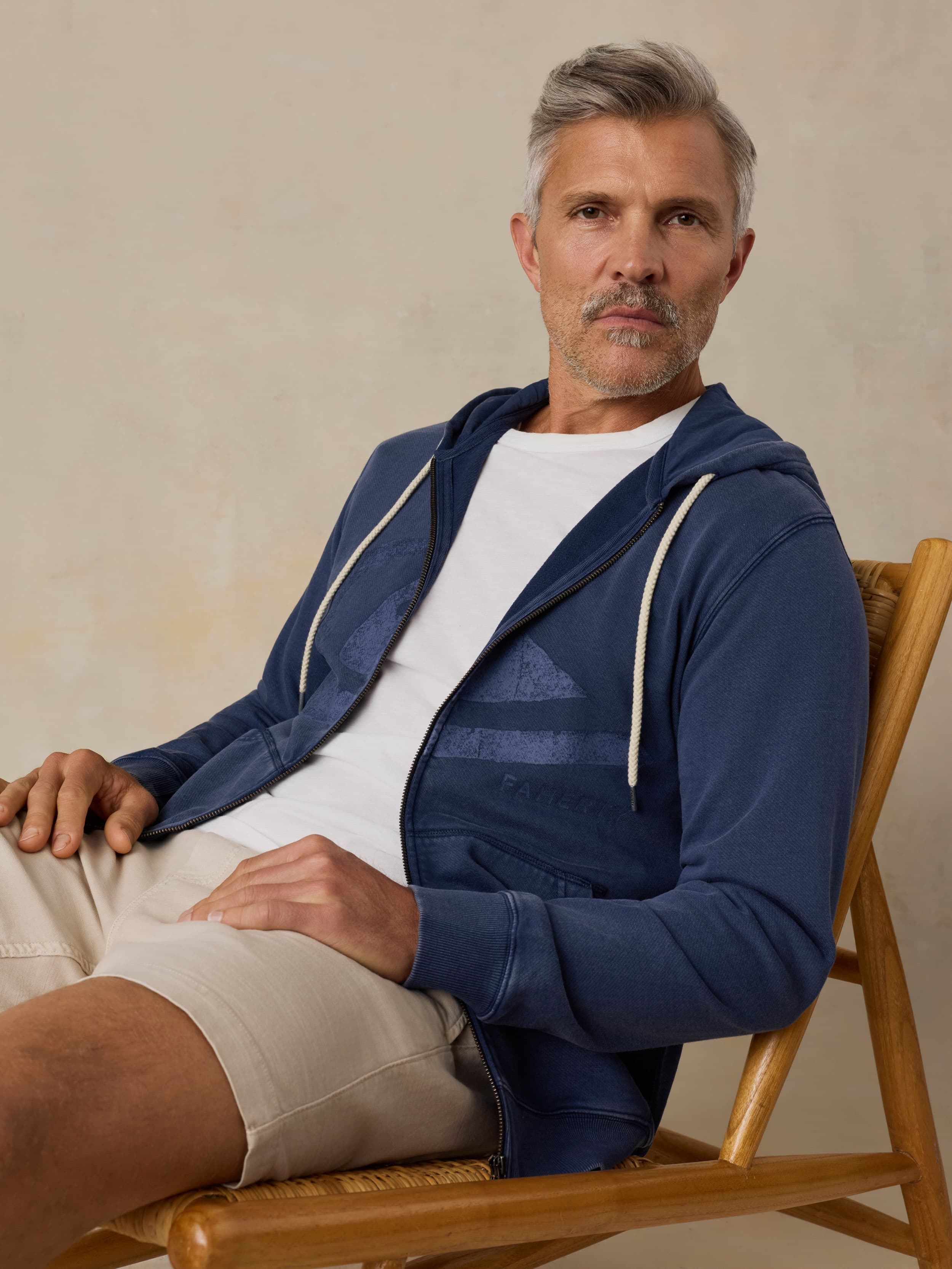 Man sitting on a wooden chair wearing a blue hoodie with a Faherty x Quiksilver and beige shorts against a beige background