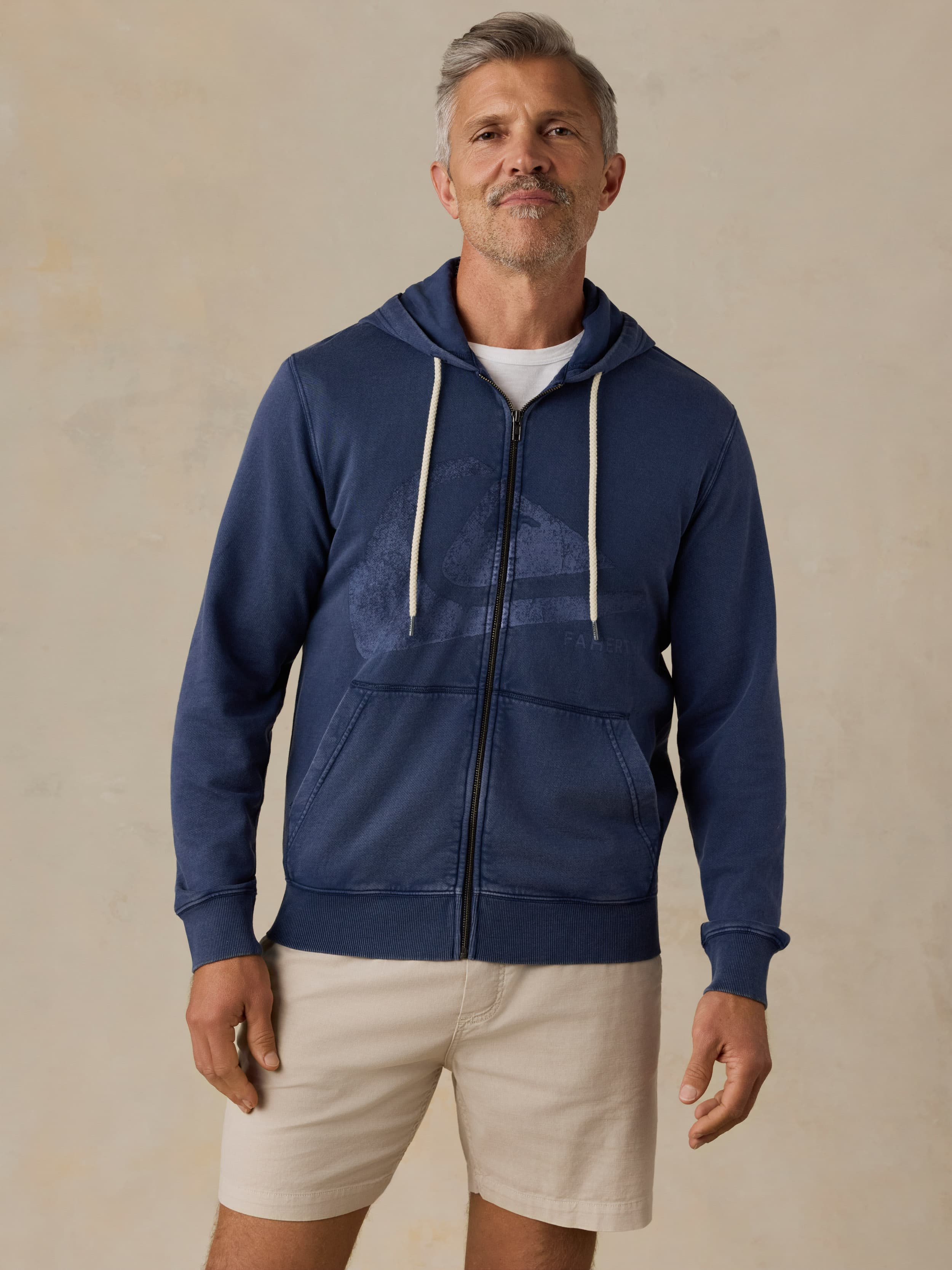 Man wearing a blue hoodie with a Faherty x Quiksilver and beige shorts against a beige background