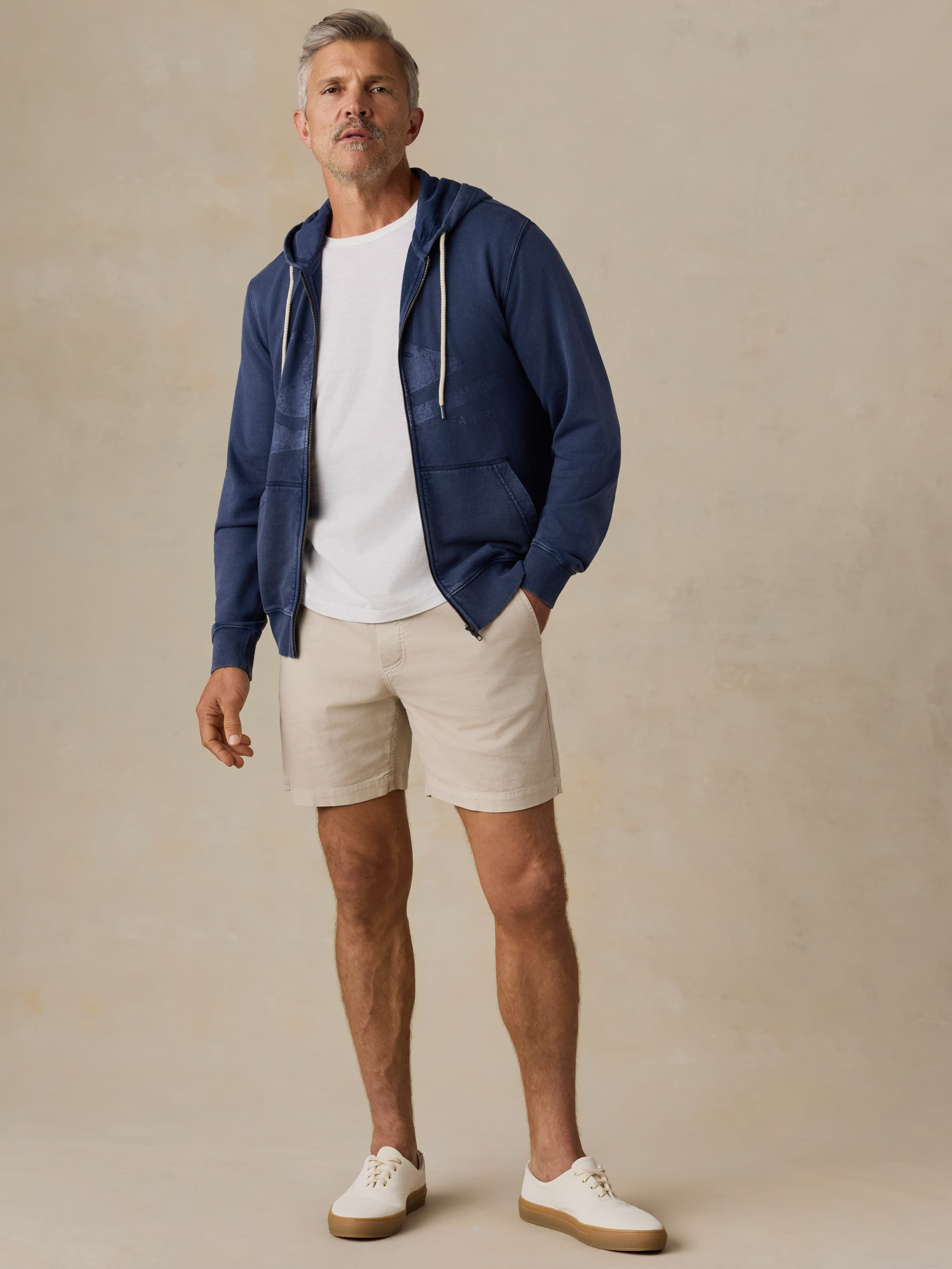 Man wearing a navy sweatshirt with Faherty x Quiksilver logo, white shirt, beige shorts, and white shoes on a beige background