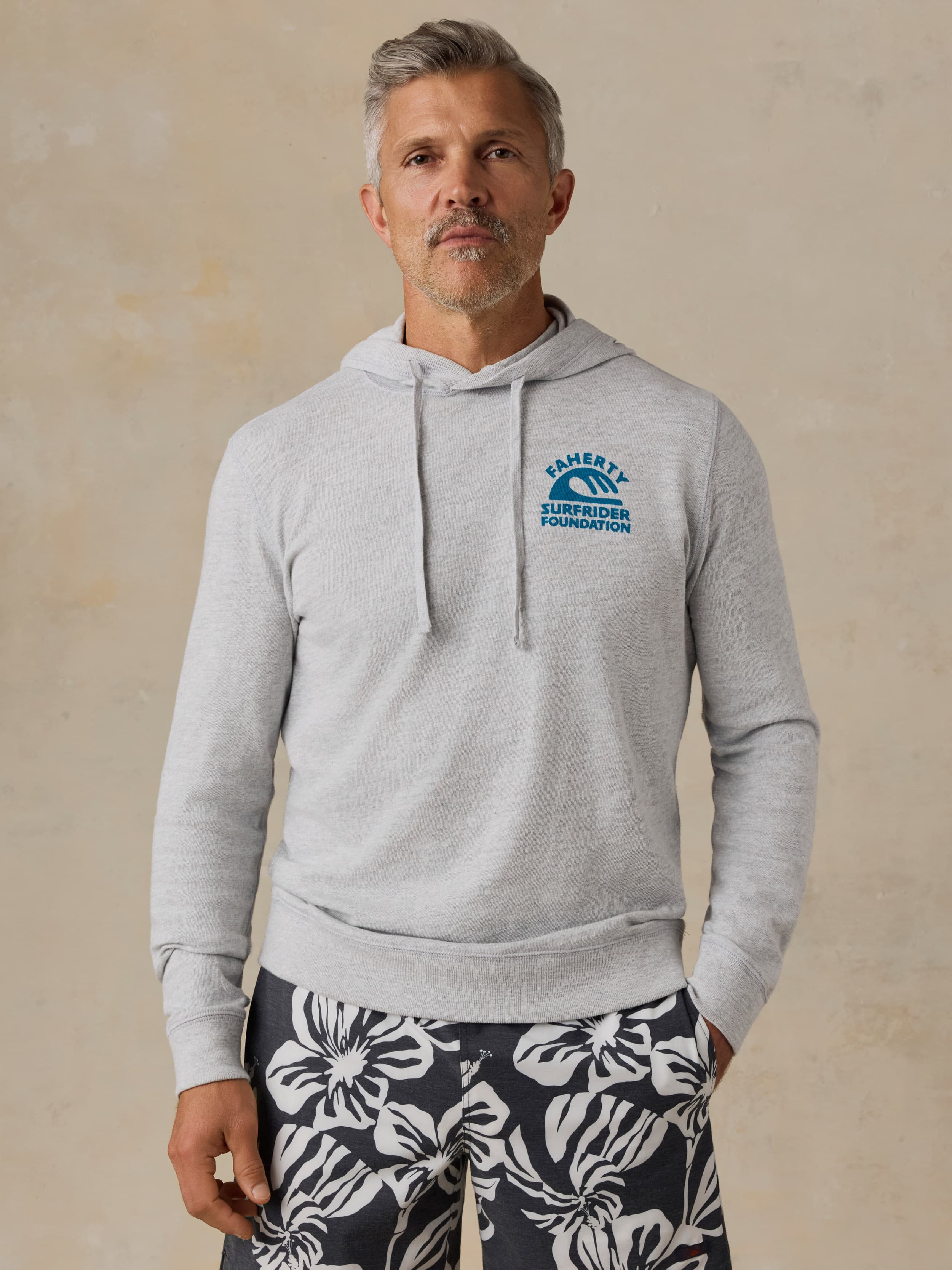 Man wearing a gray hoodie with a Faherty x Surfrider Foundation logo on a beige background