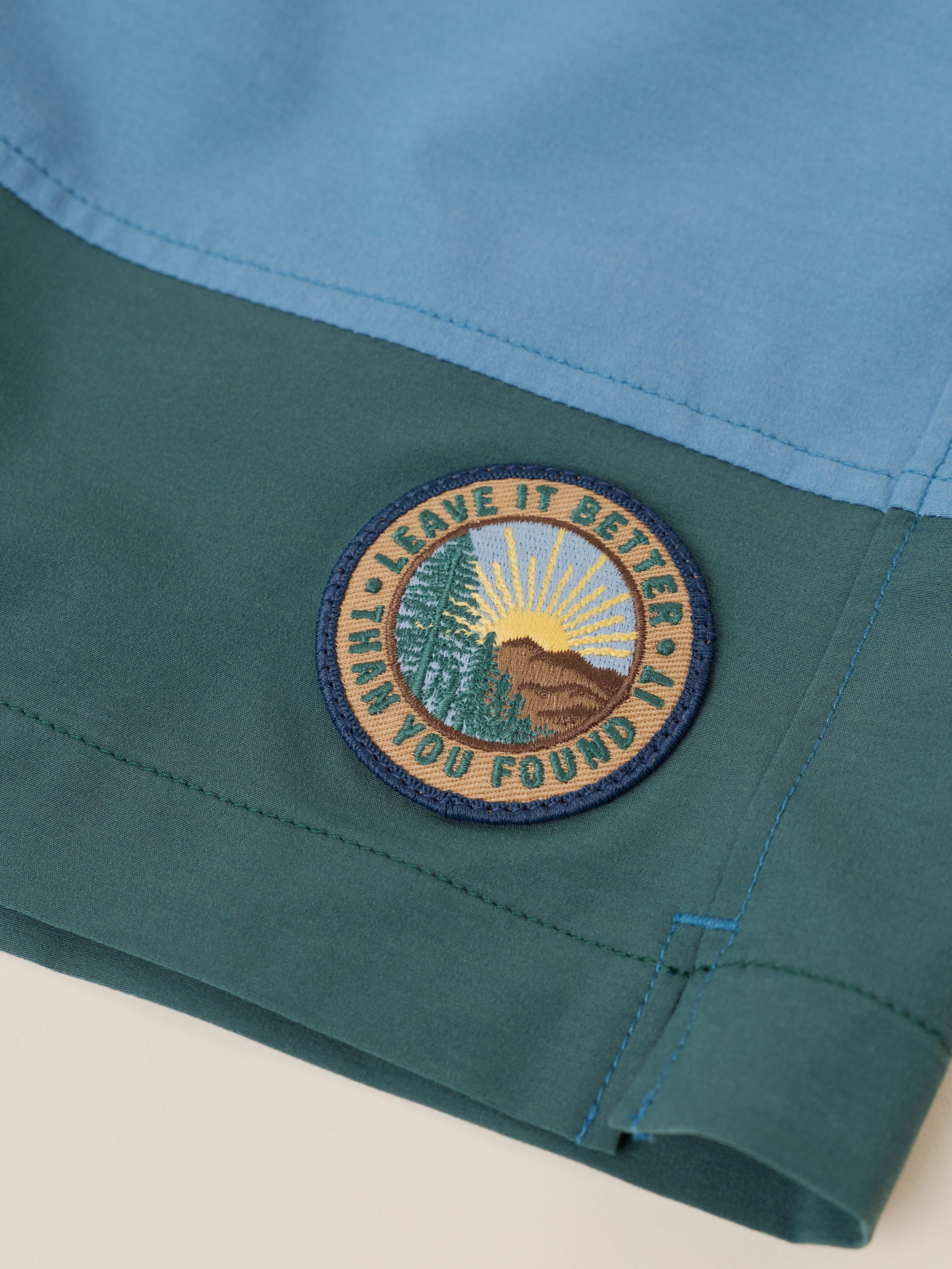 Close-up of a folded garment showing a color-blocked design with light blue on top and teal green on the bottom. A circular embroidered patch reading 'LEAVE IT BETTER THAN YOU FOUND IT' with a mountain, trees, and sun is stitched on the teal section.