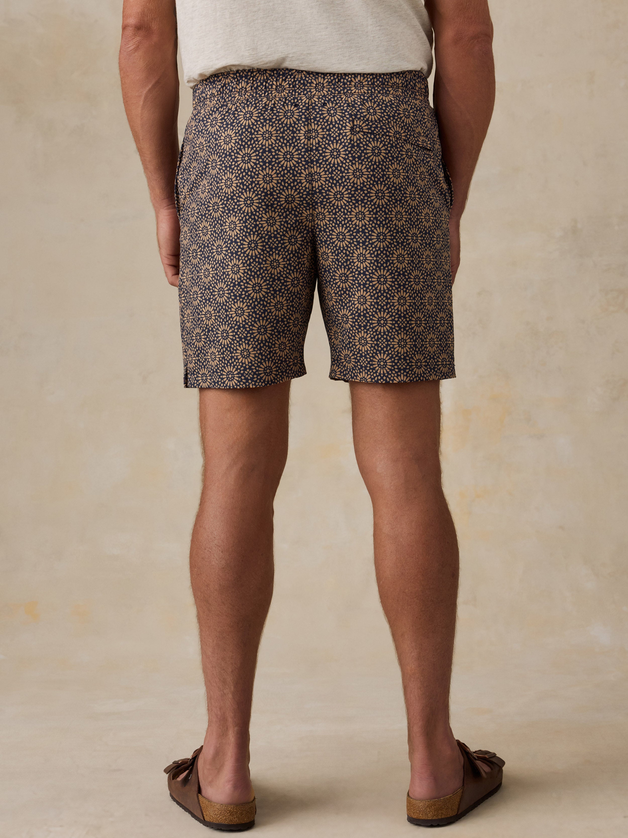 Man wearing a light colored t-shirt, brown patterned swim shorts, and sandals against a beige background