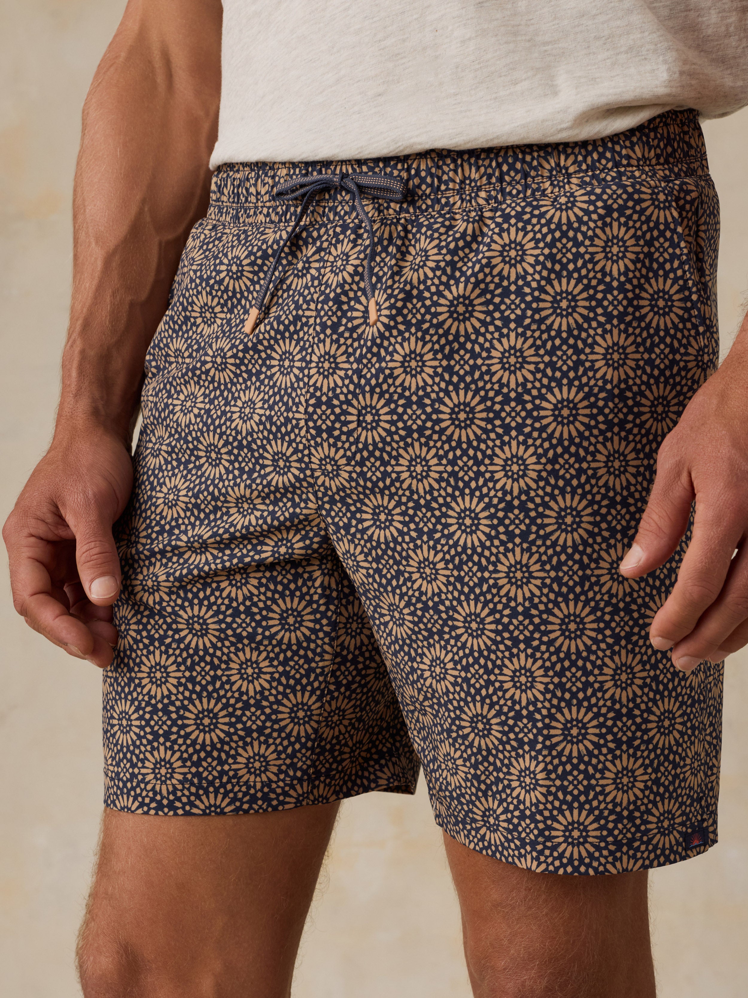 Man wearing a light colored t-shirt, brown patterned swim shorts, and sandals against a beige background