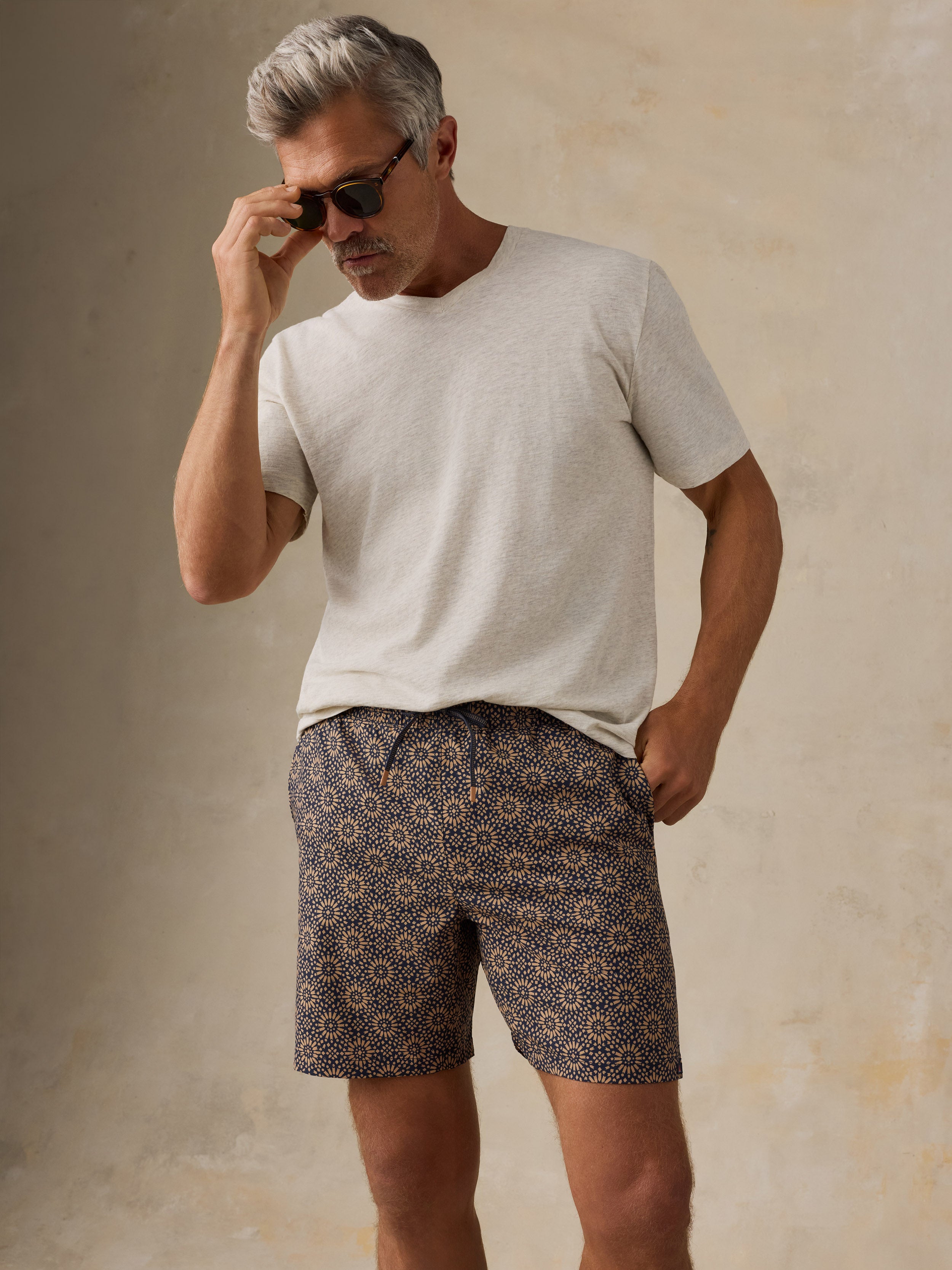 Man wearing a light colored t-shirt, brown patterned swim shorts, and sandals against a beige background