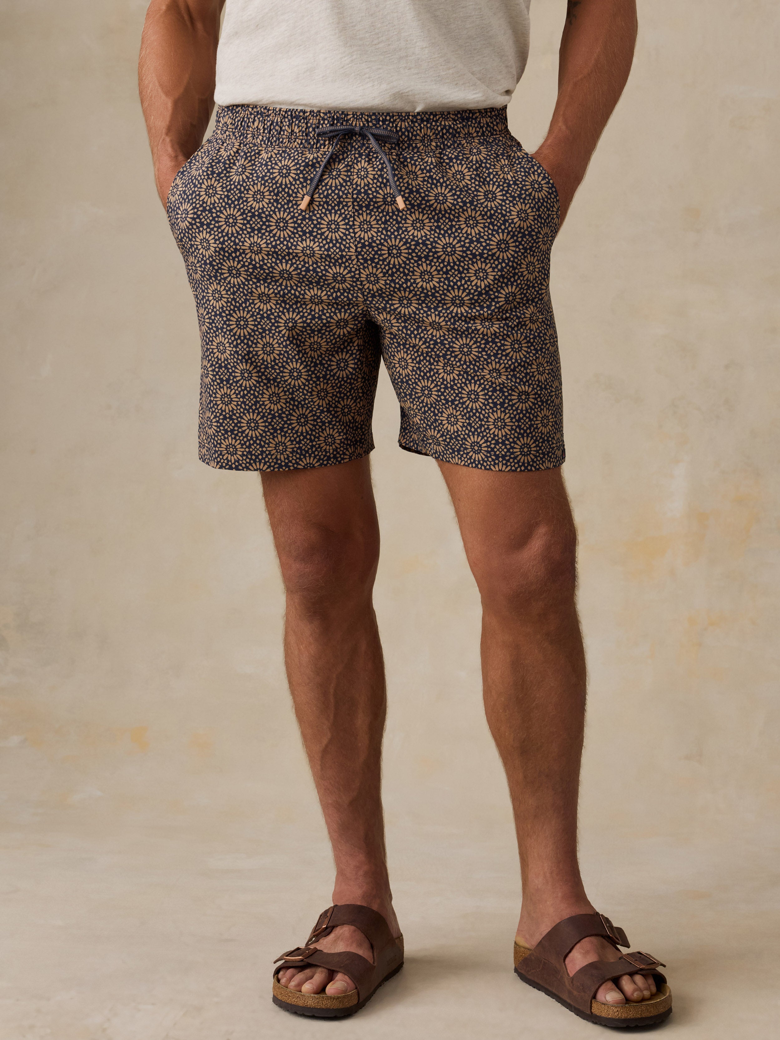 Man wearing brown patterned swim shorts and sandals against a beige background