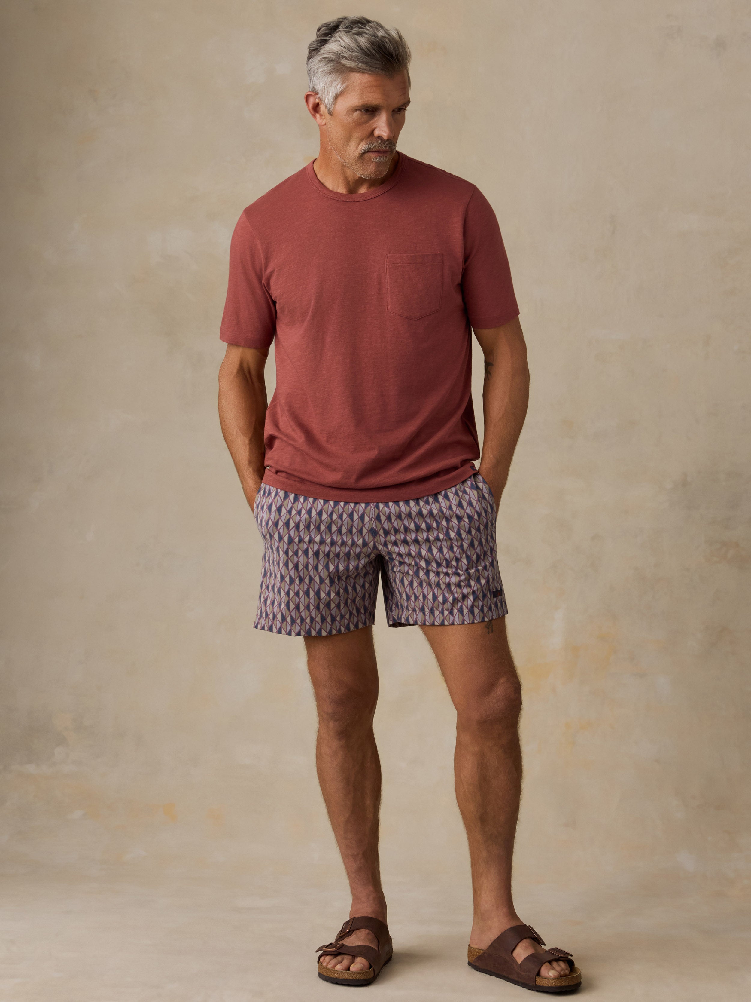 Man wearing a red t-shirt, purple geometric print swim shorts, and sandals against a beige background
