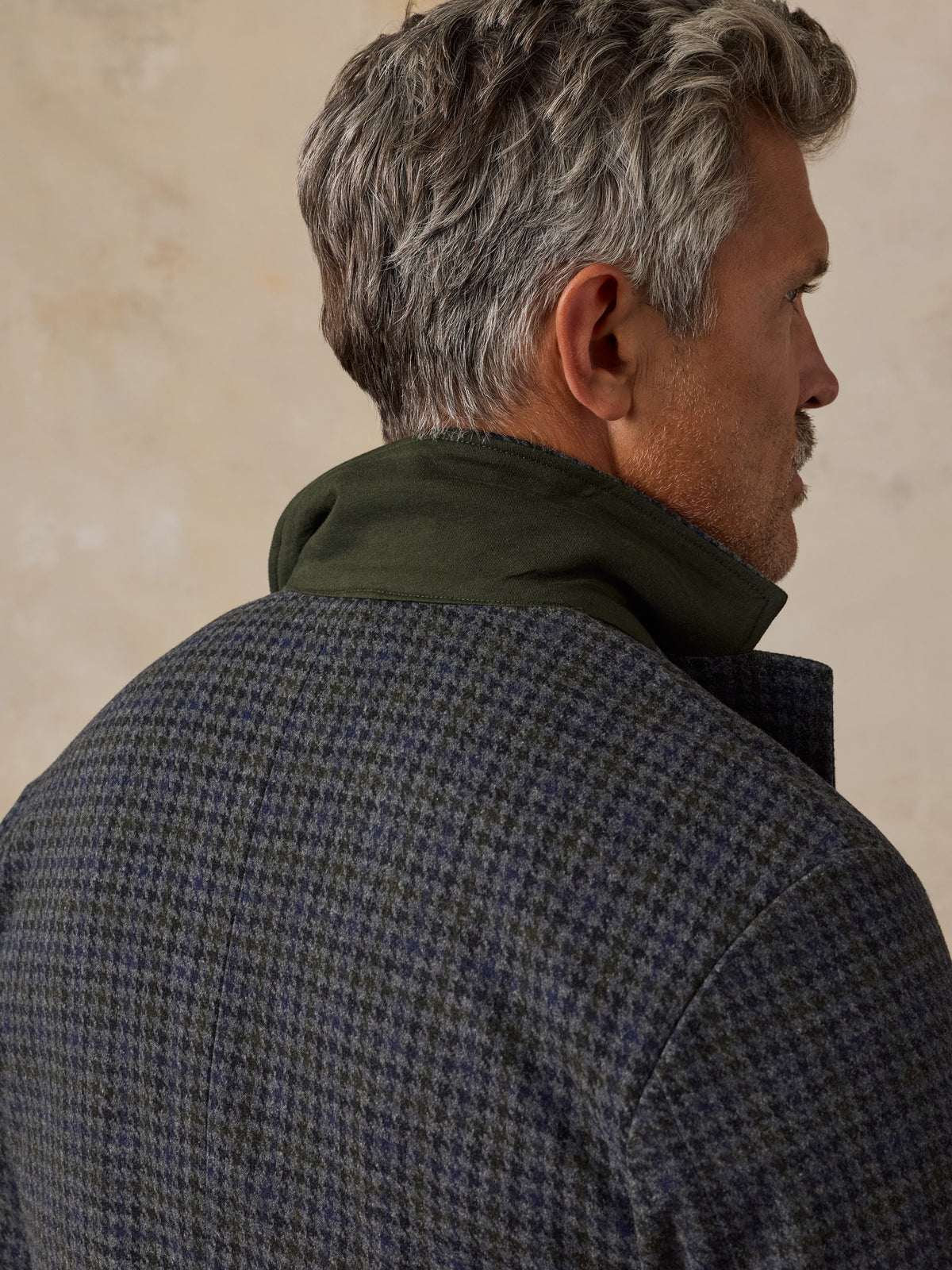 A close-up side profile of a man wearing a textured blue and gray houndstooth jacket with a dark olive green collar, set against a neutral beige background.