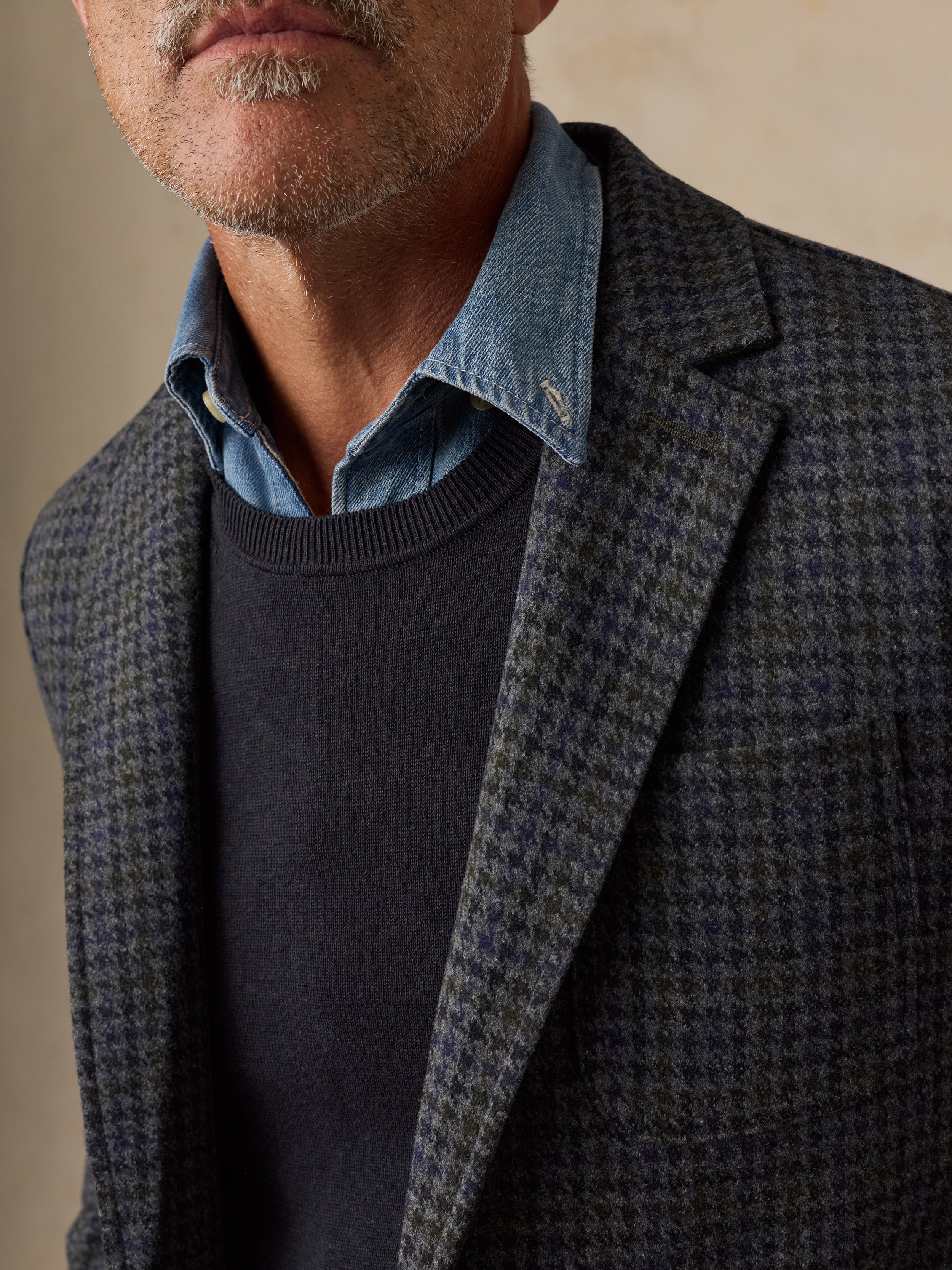 Close-up of a man wearing a dark blue and gray houndstooth blazer over a navy sweater and a light blue chambray shirt collar, against a warm beige background.
