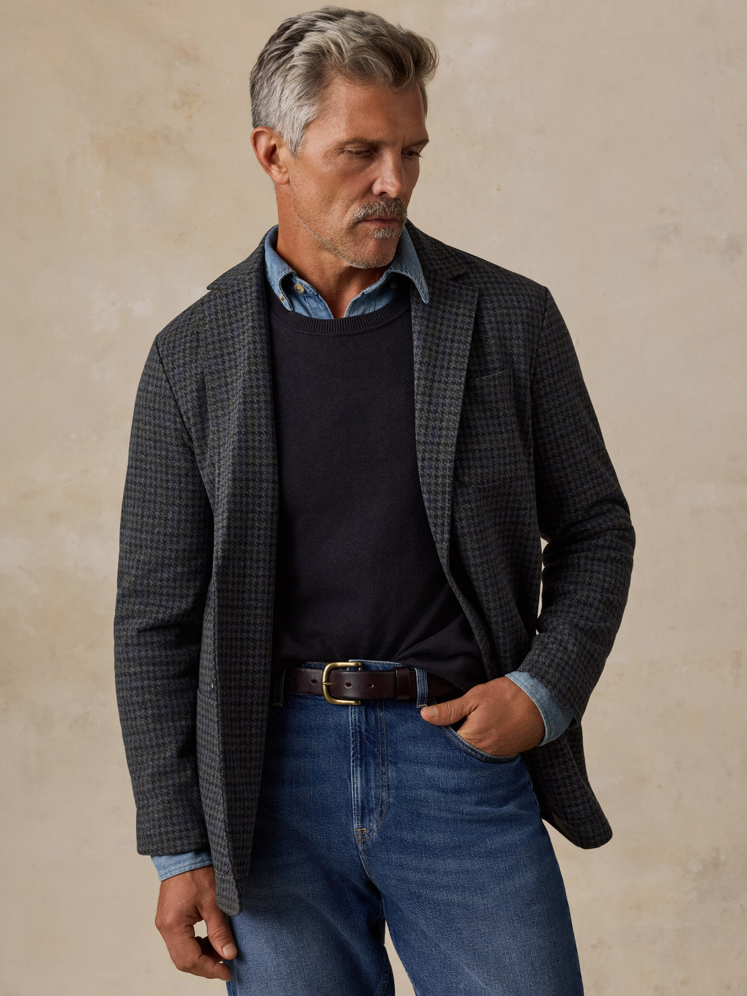 A man wearing a dark gray and black checkered blazer over a black sweater, blue collared shirt, and blue jeans, standing against a textured beige background.