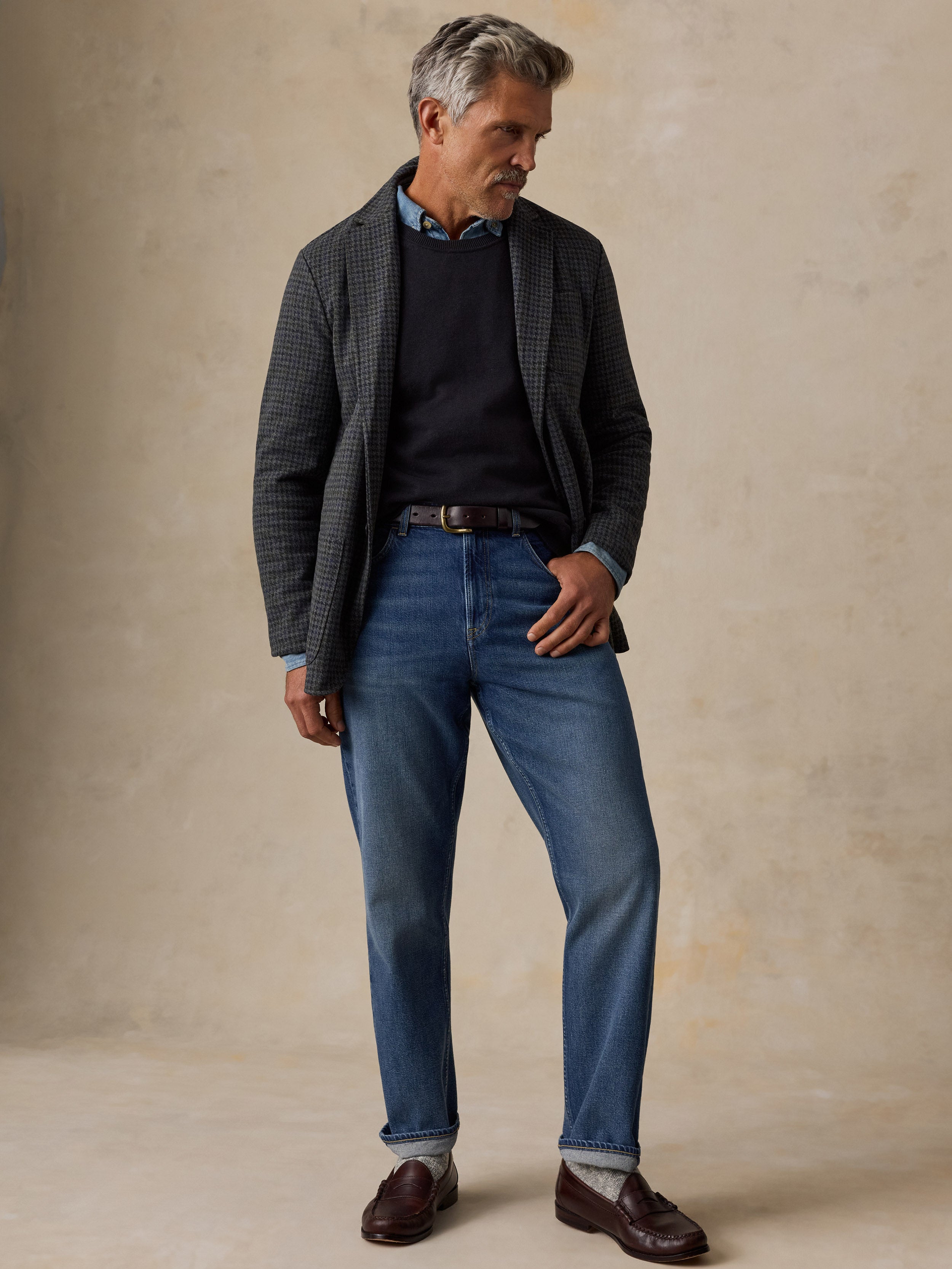 A man wearing a dark gray patterned blazer, navy sweater, blue jeans, and brown leather loafers, standing against a textured beige studio backdrop.