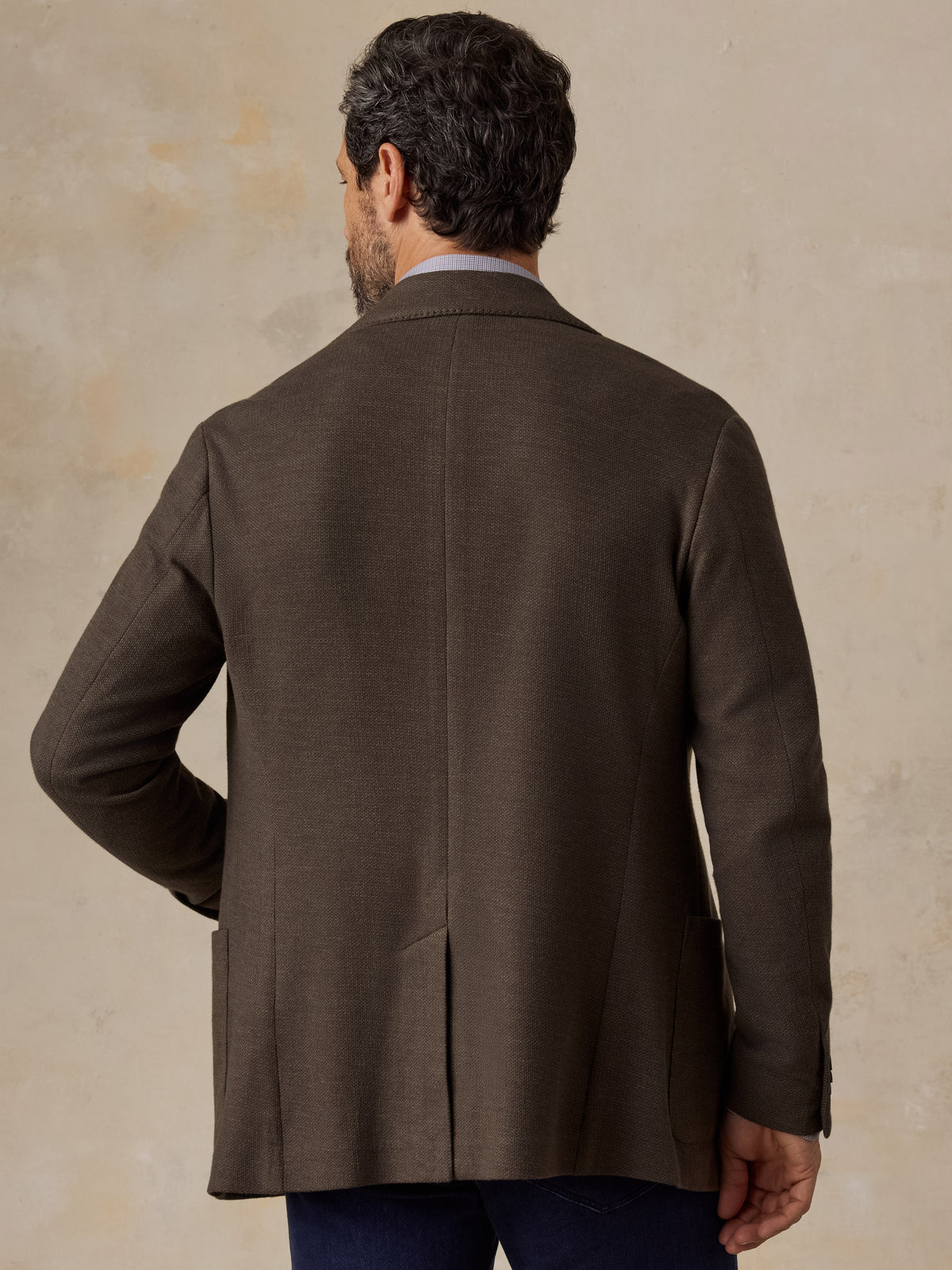 A man viewed from behind wearing a brown blazer over a blue shirt and blue jeans, image 2 of 5