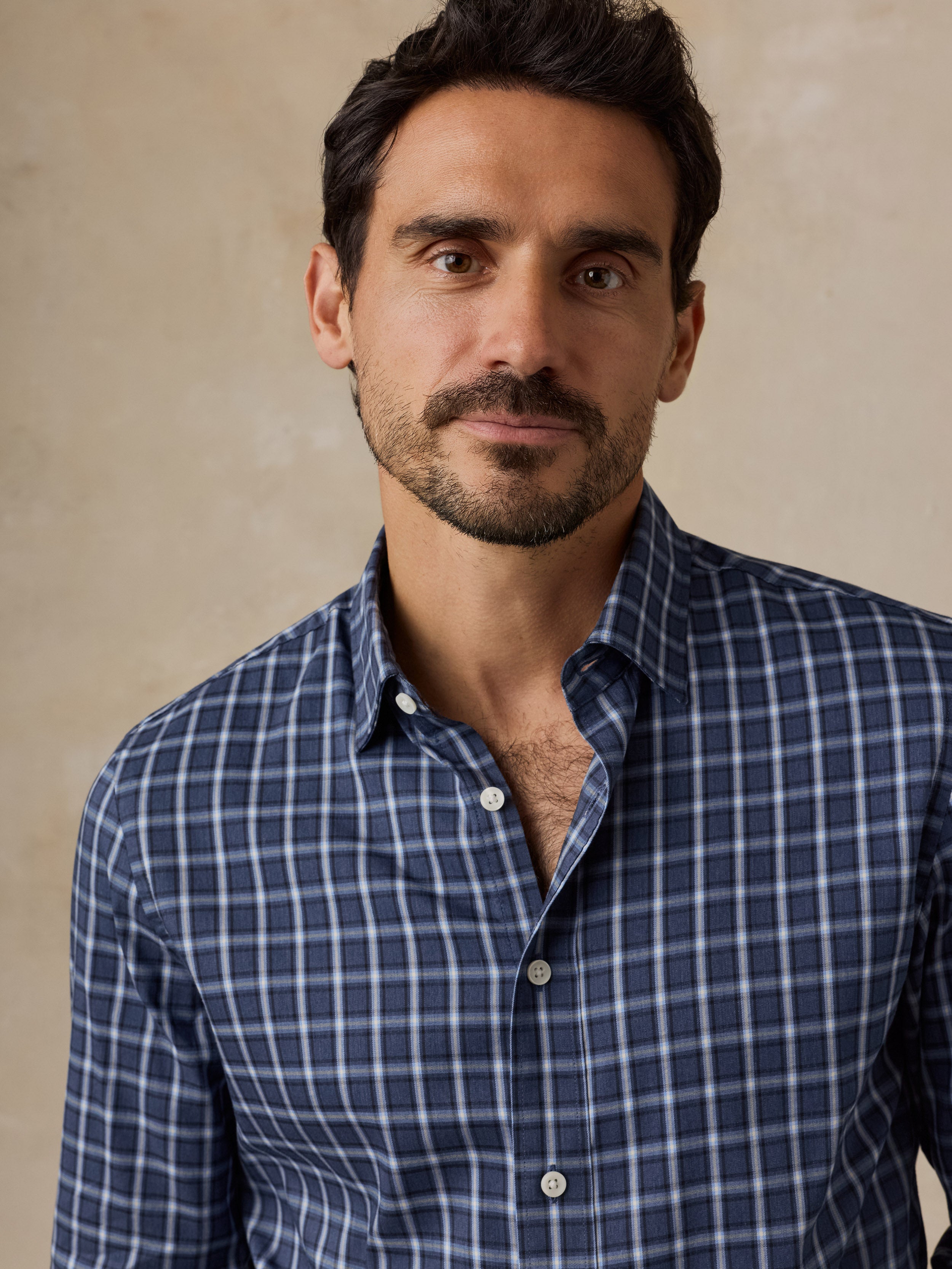 A man wearing a blue and white checkered button-down shirt, photographed from the chest up against a neutral beige background.