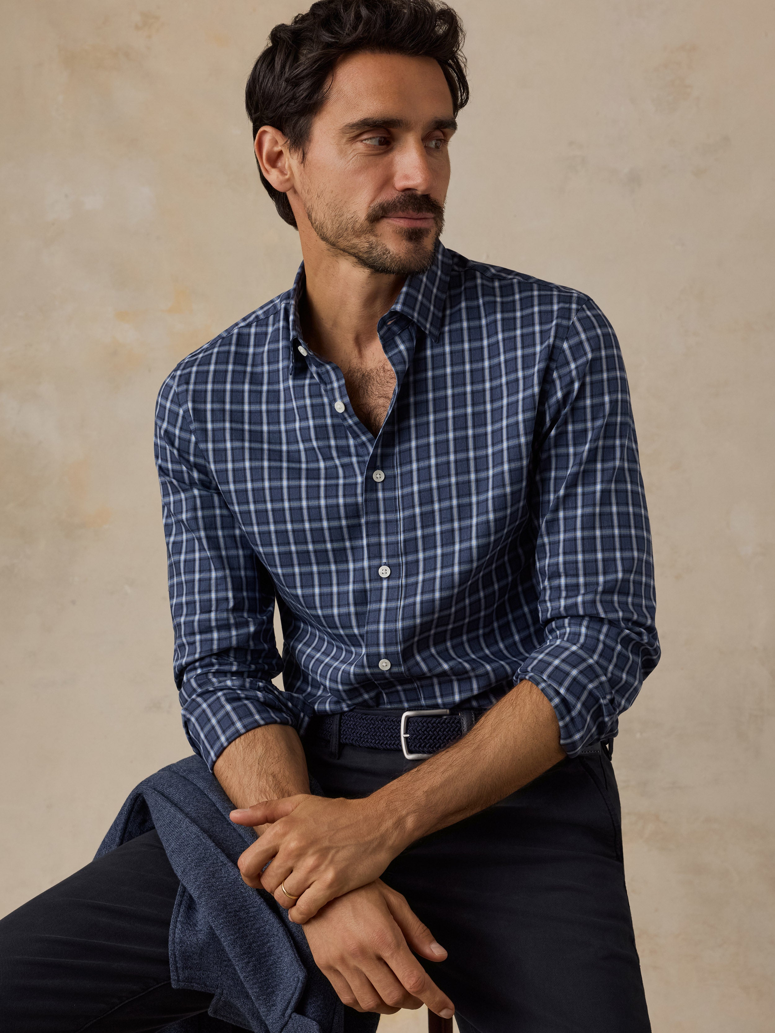 A man wearing a navy blue and white checkered long-sleeve button-down shirt, paired with dark trousers and a belt, sitting against a textured beige background.