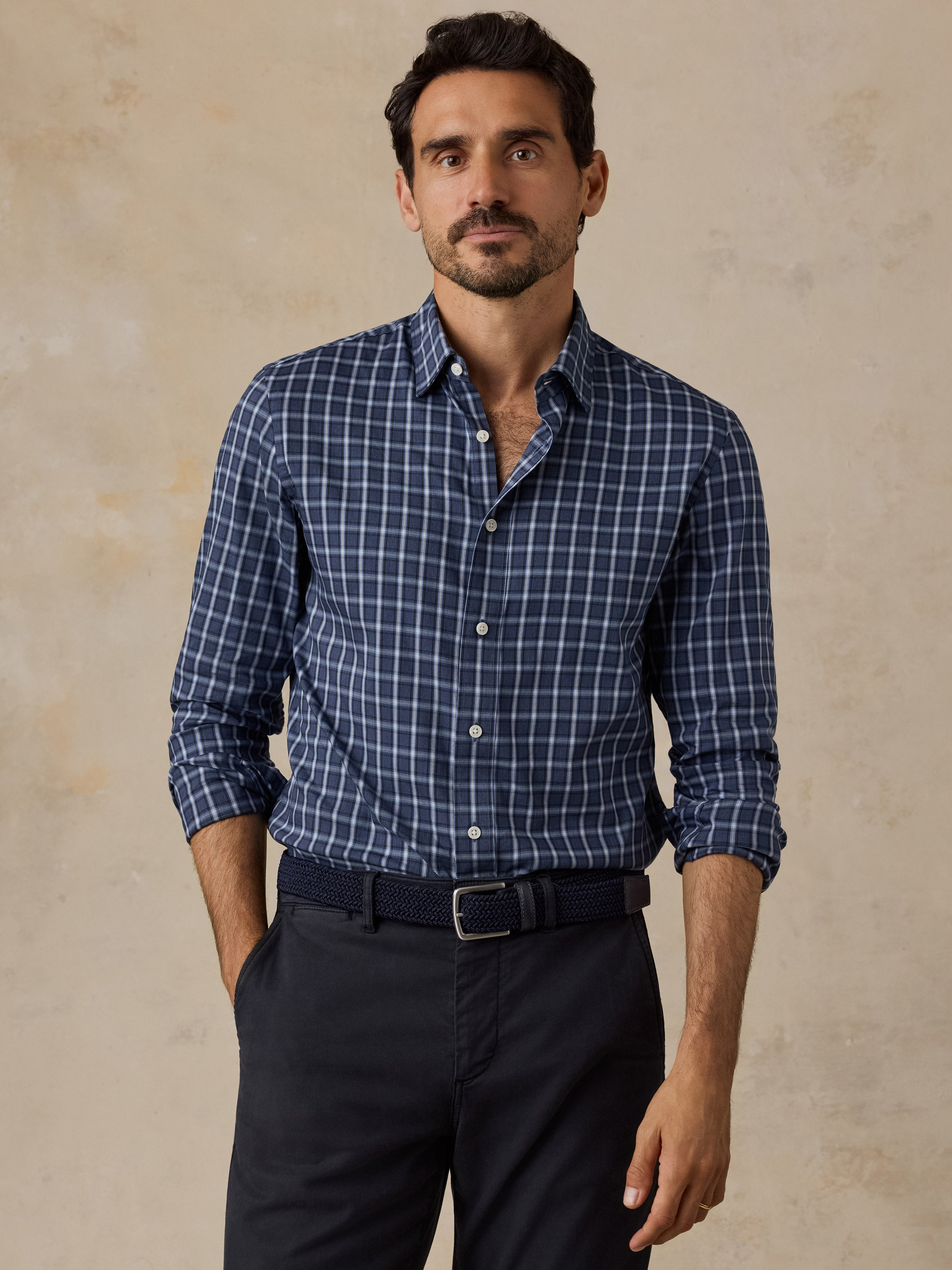 A man wearing a navy blue and white checkered long-sleeve button-down shirt, paired with dark trousers and a dark belt, standing against a textured beige background.