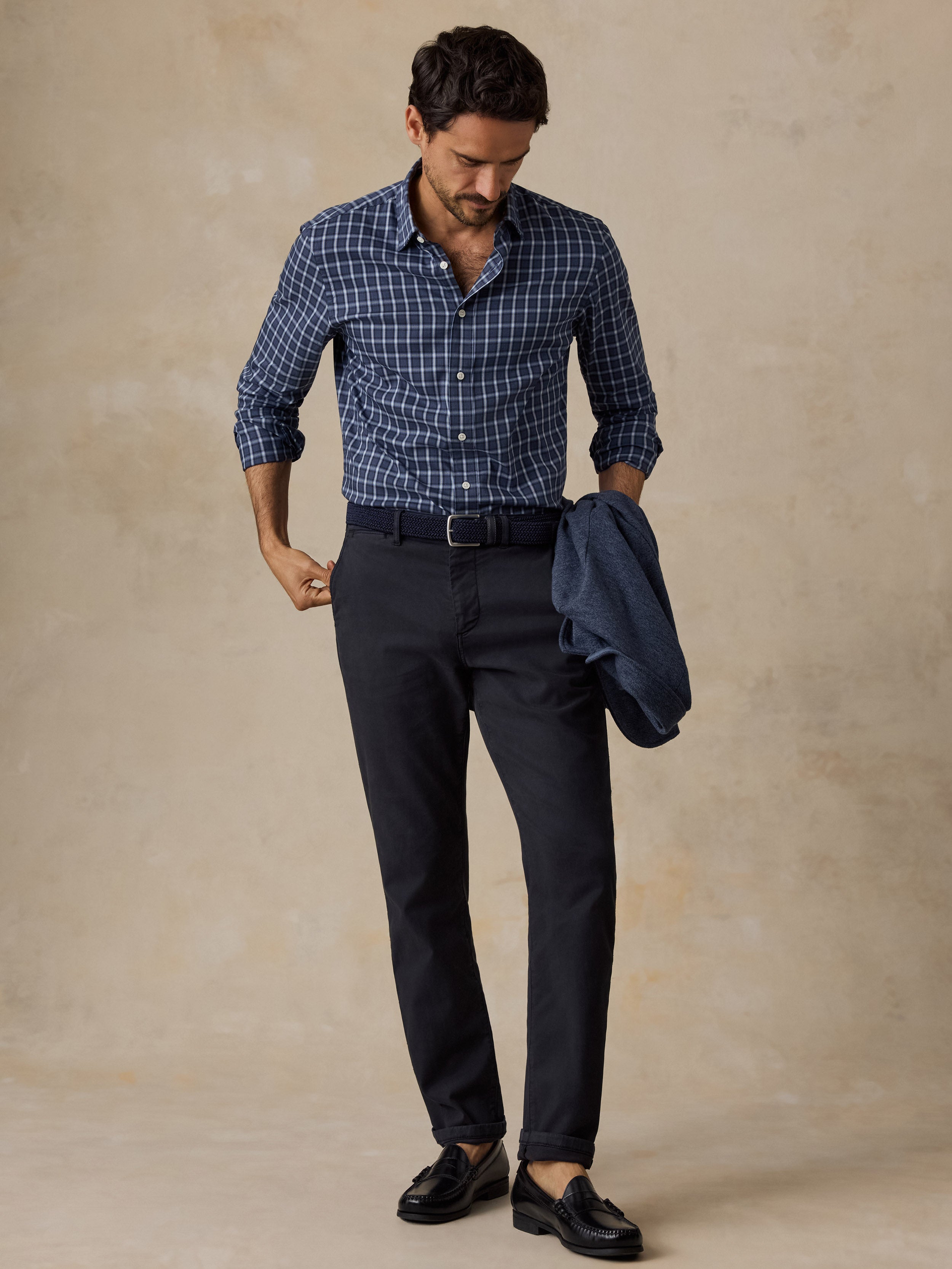 A man modeling a blue and white checkered long-sleeve button-down shirt, dark navy trousers, and black loafers, standing against a textured beige studio backdrop.