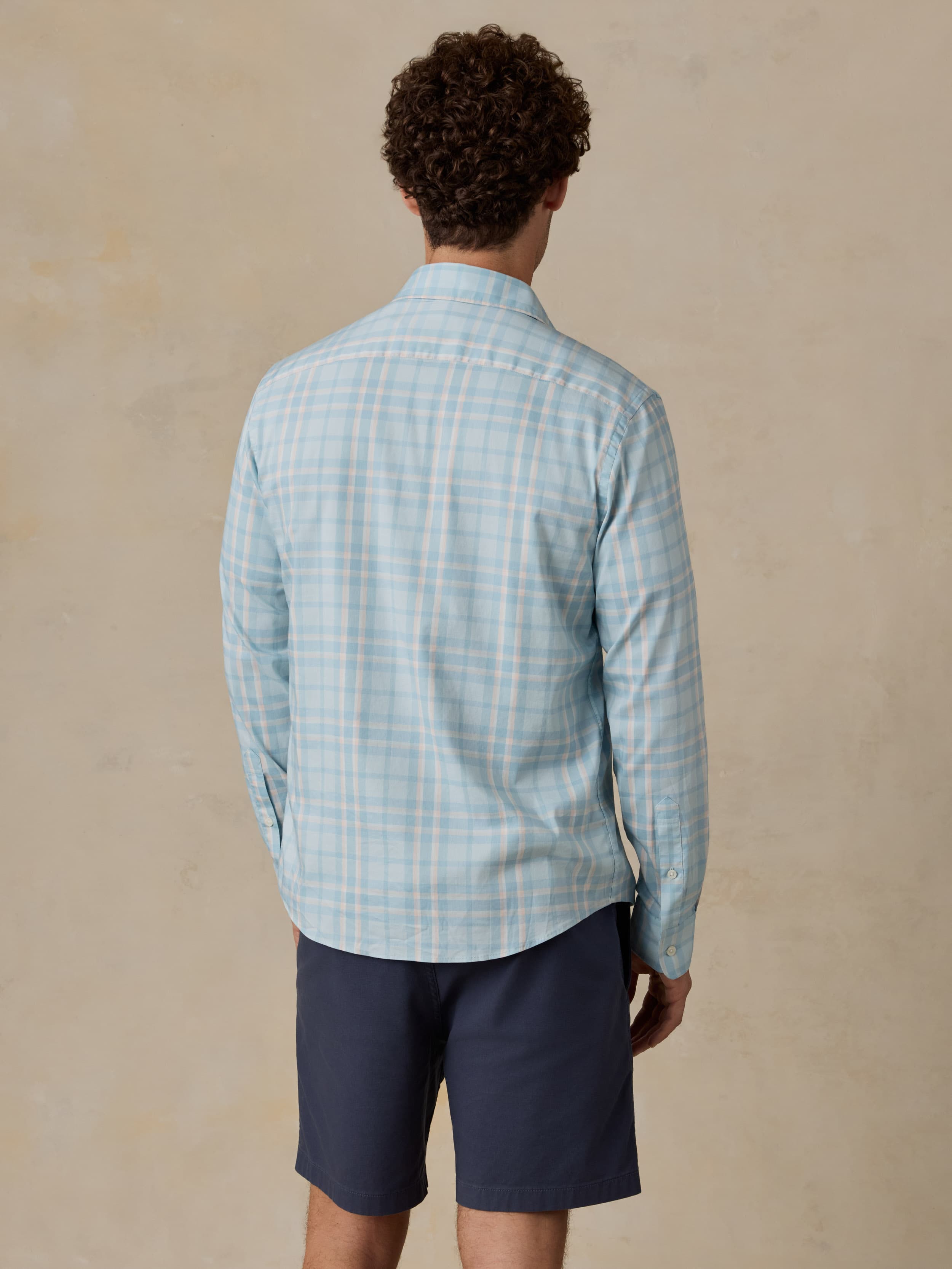 Man wearing blue and white plaid shirt with navy shorts against a beige background