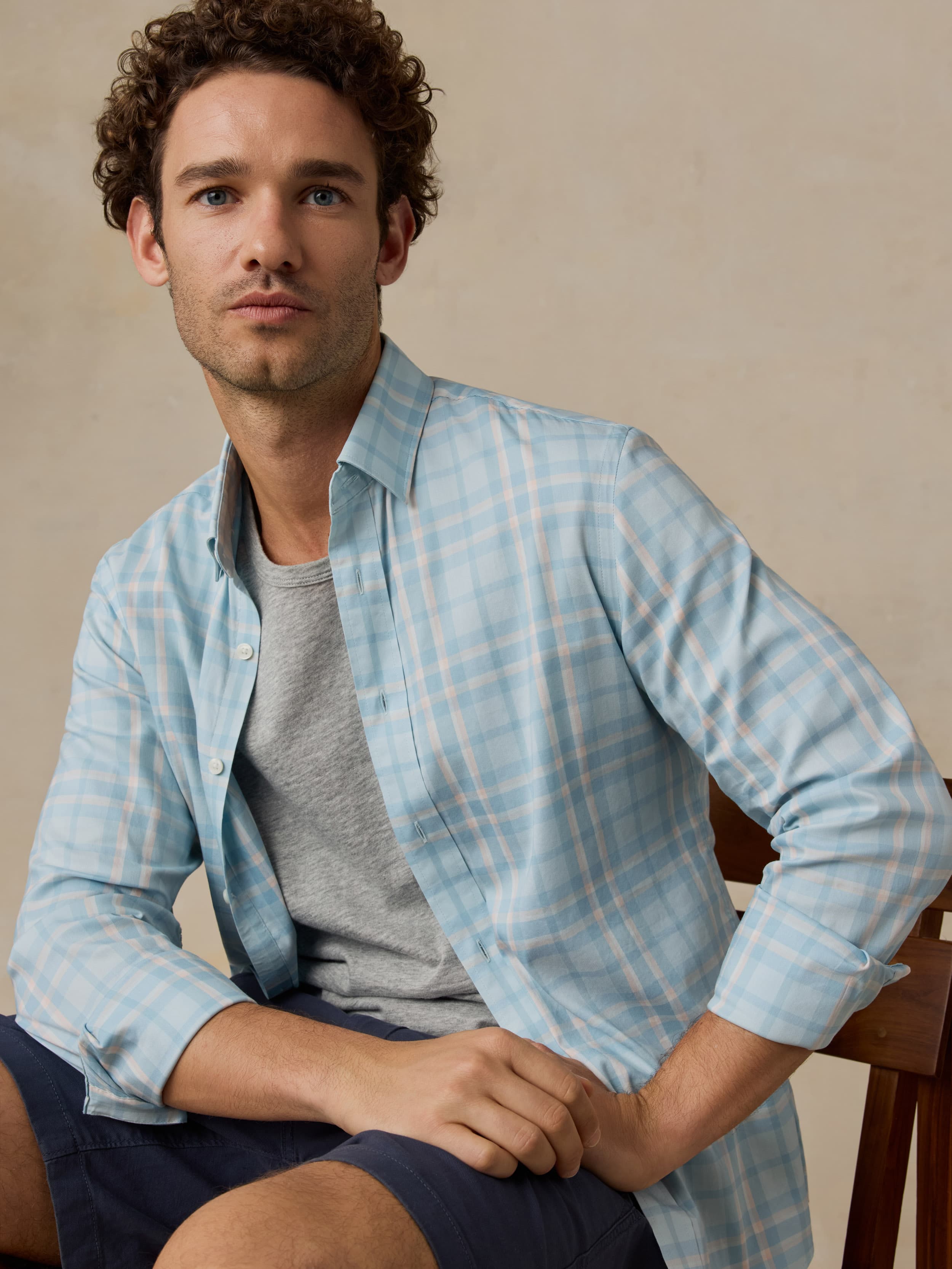 Man sitting on a wooden chair wearing blue and white plaid shirt with navy shorts against a beige background