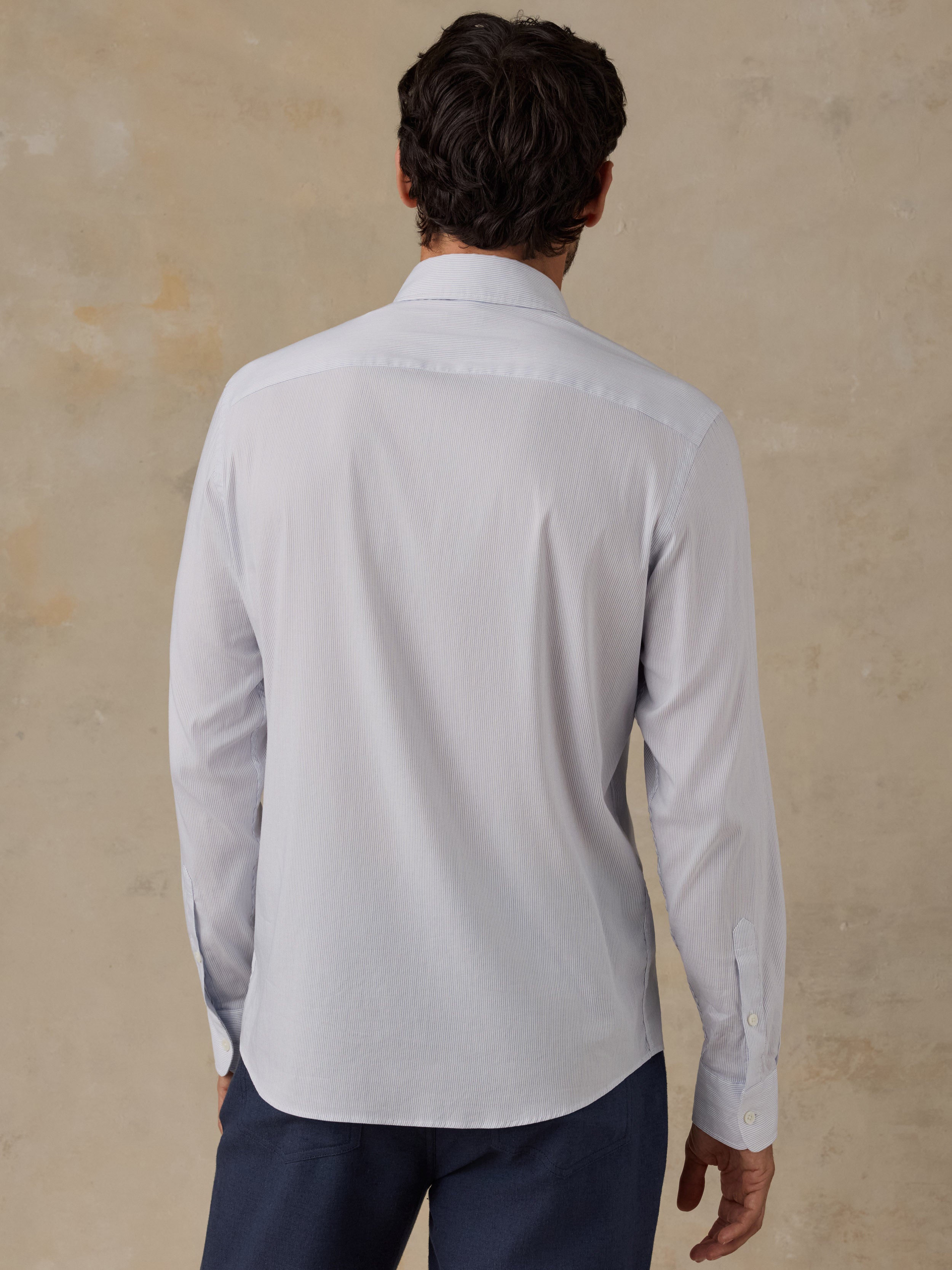 A man viewed from behind wearing a light gray long-sleeve button-down shirt and navy trousers, standing against a textured beige wall.