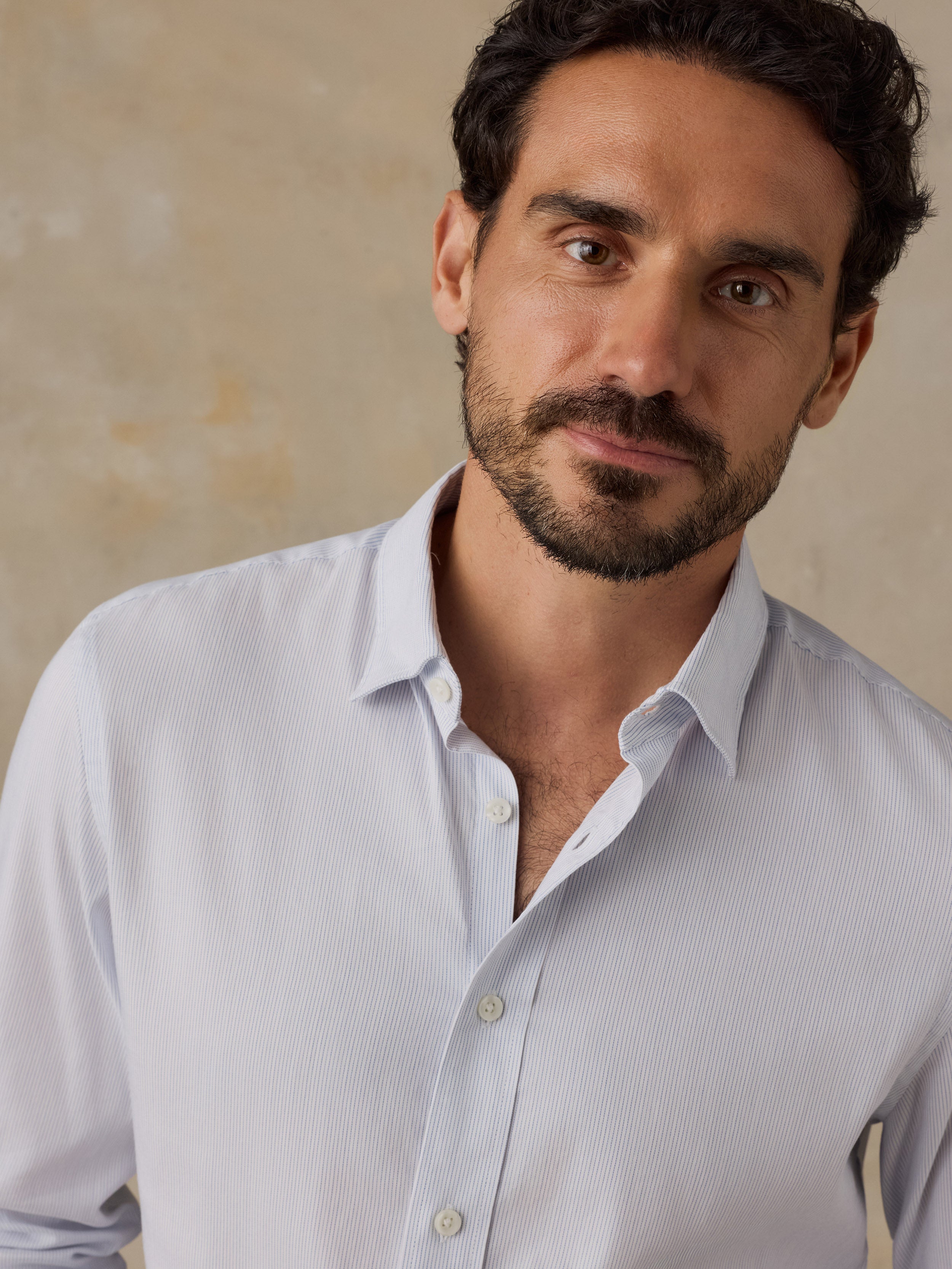A close-up portrait of a man wearing a light blue long-sleeve button-down shirt against a textured beige background.