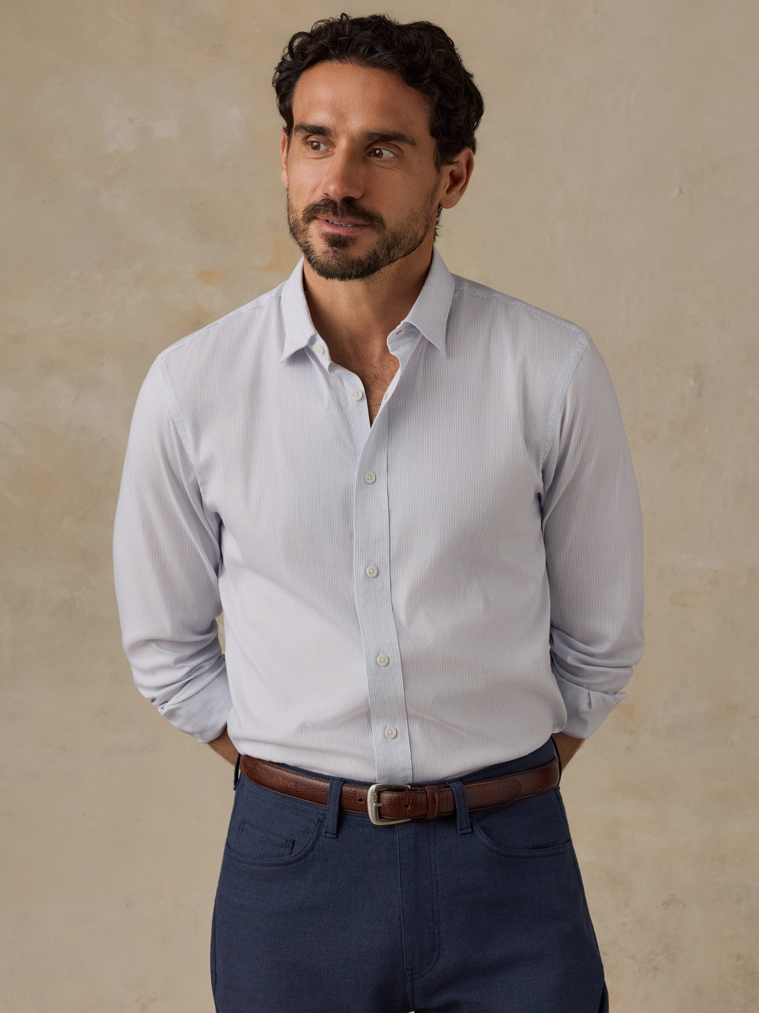 A man wearing a light blue long-sleeve button-down dress shirt, navy blue trousers, and a brown leather belt, standing against a textured beige studio backdrop.