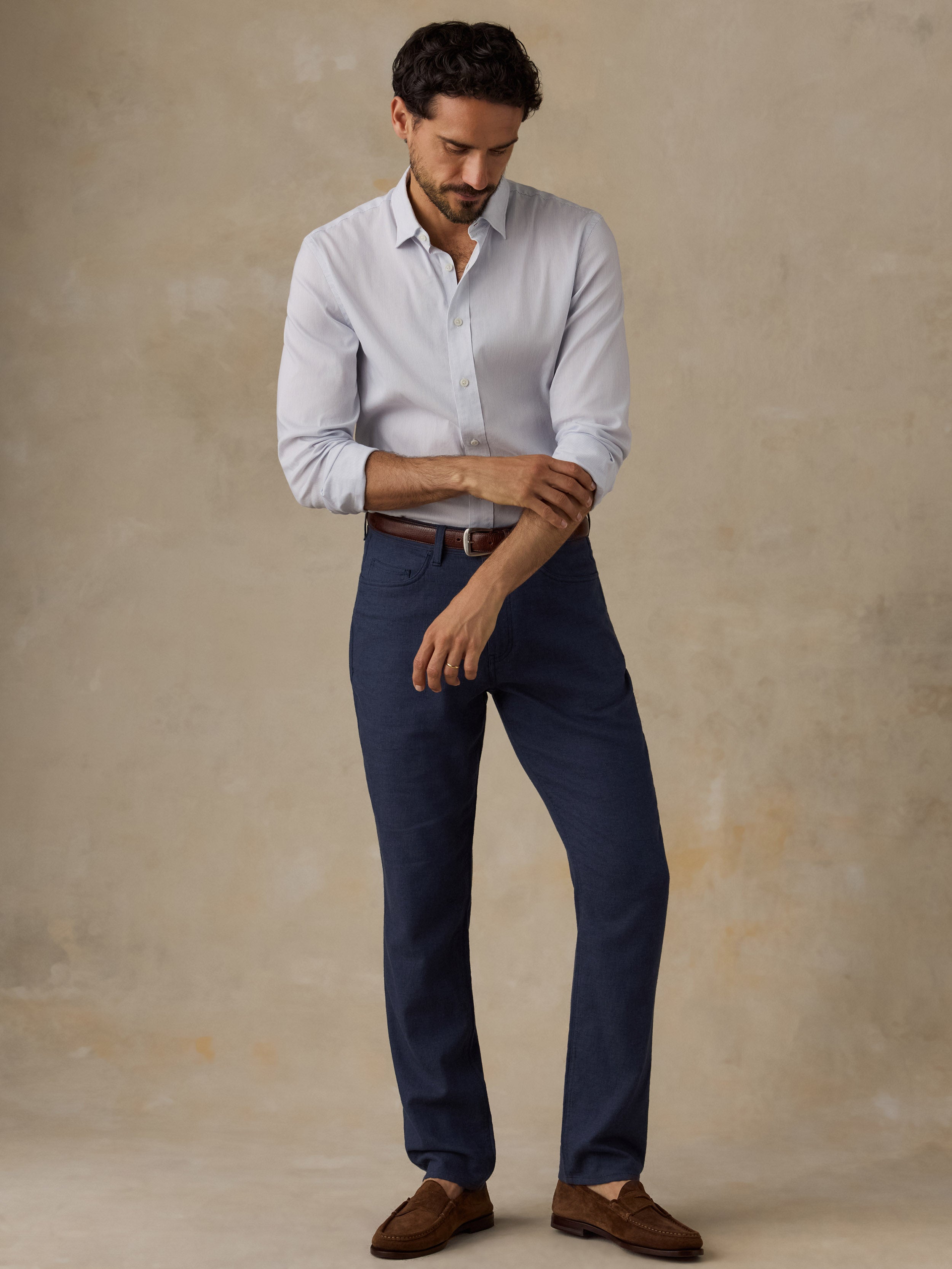 A man wearing a light blue long-sleeve button-down shirt, navy blue trousers, a brown belt, and brown leather loafers, standing against a textured beige studio backdrop.