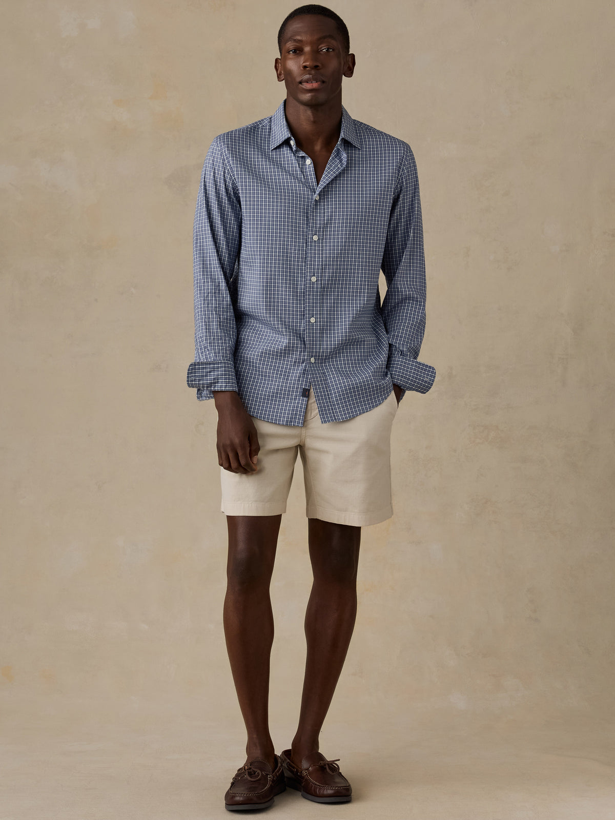 A man wearing a blue checkered shirt, beige shorts, and boat shoes, image 2 of 5