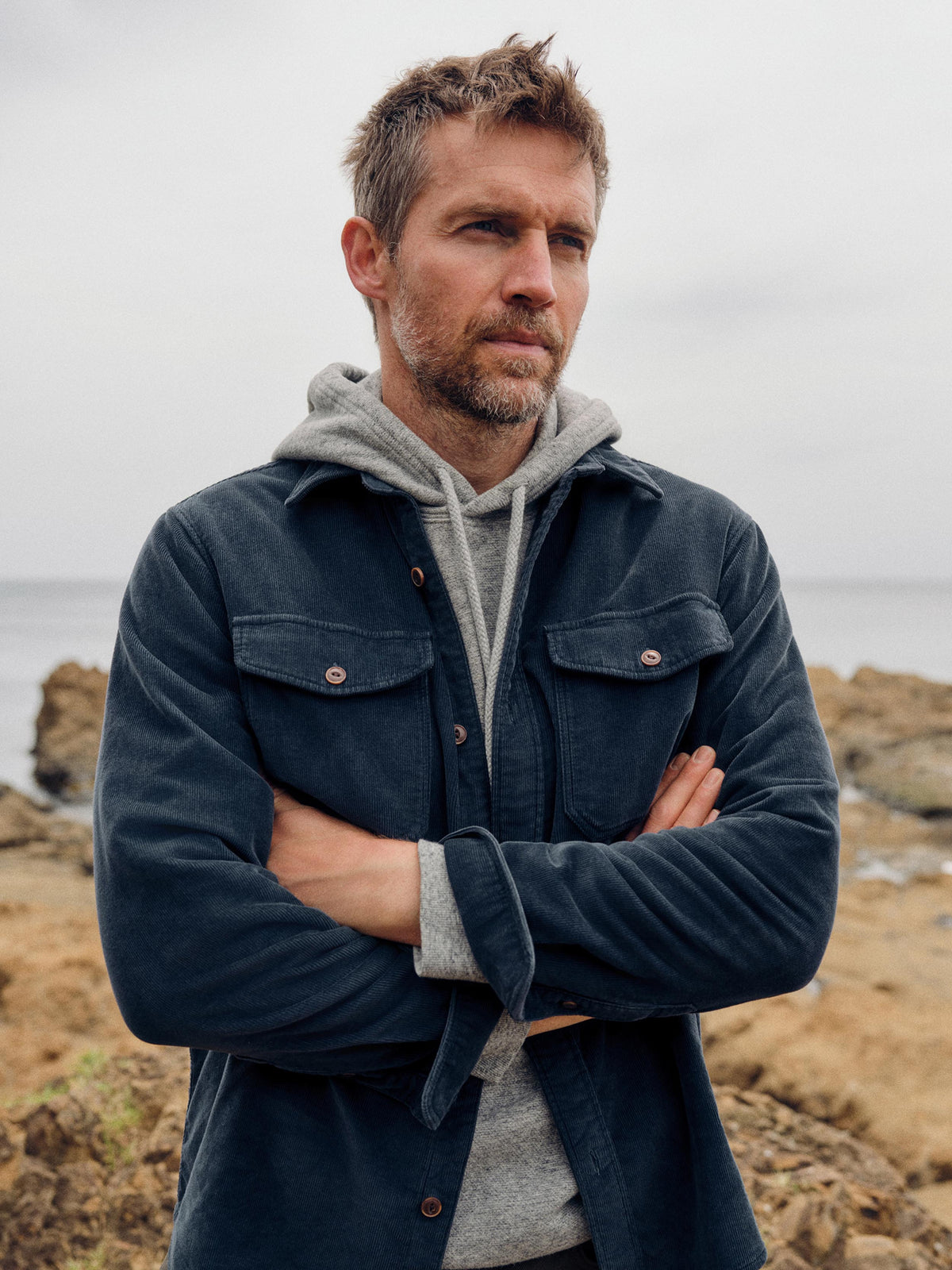 A man standing outdoors on a rocky shoreline with his arms crossed, wearing a dark navy overshirt over a light gray hoodie., image 2 of 6
