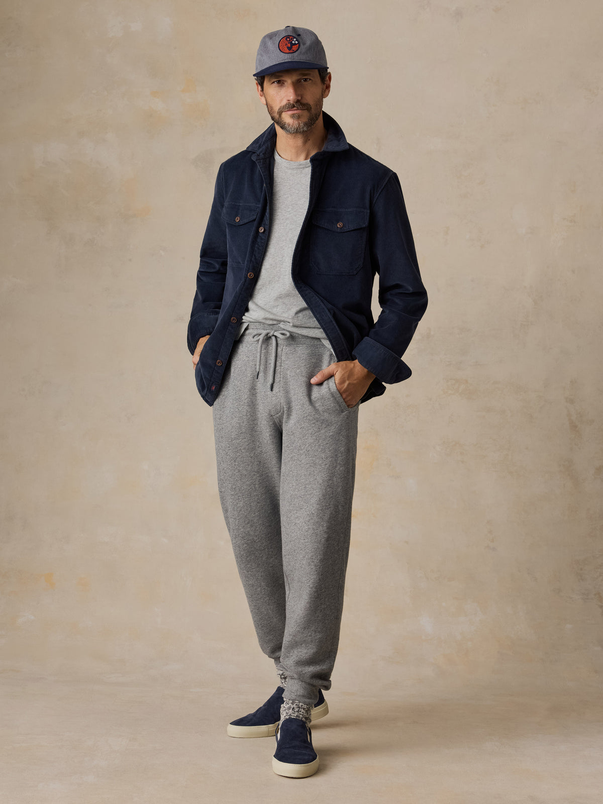 A man wearing a navy blue overshirt with a grey tee, sweatpants, and blue sneakers, image 6 of 6