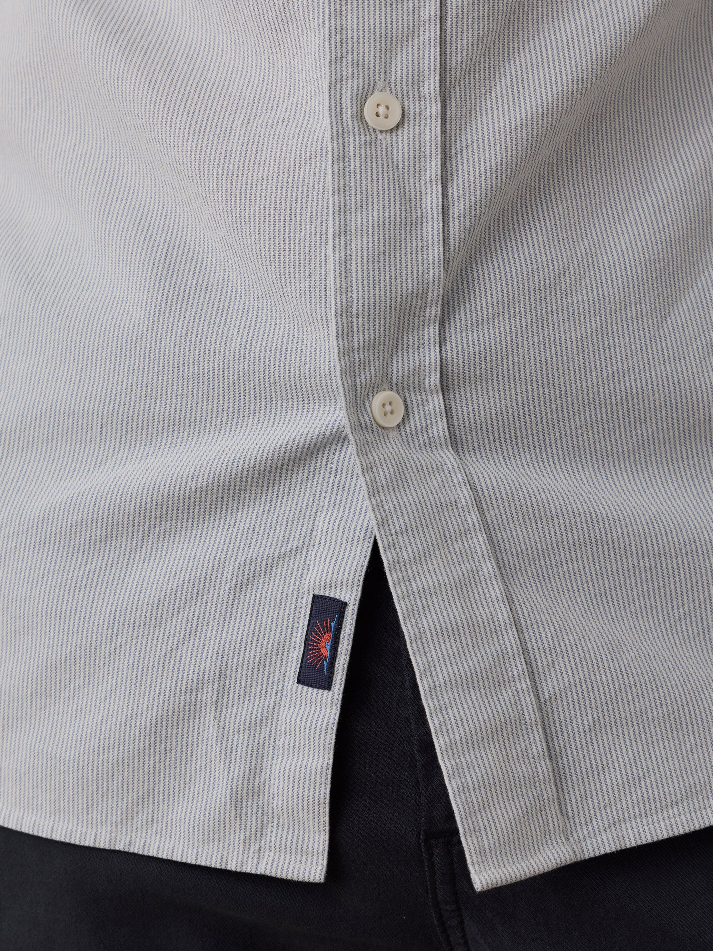 Close-up of a light blue and white striped button-down shirt with a small navy Faherty brand tag featuring a red sun logo, worn over dark pants.