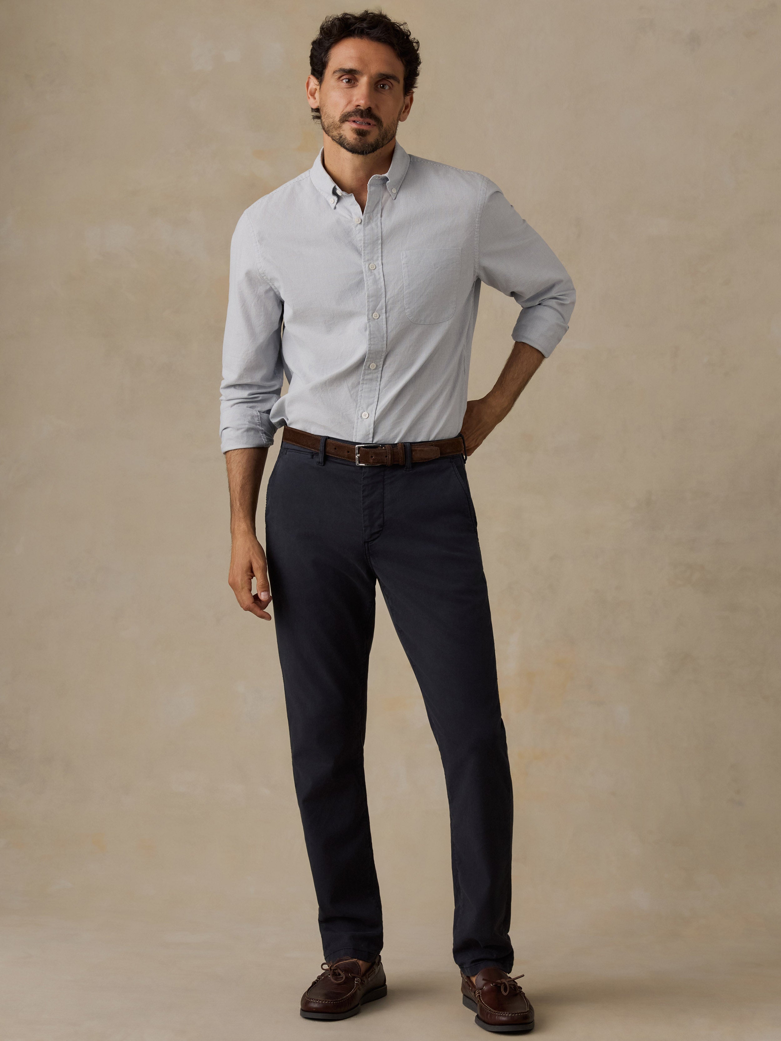 A man modeling a light blue long-sleeve button-down shirt, dark navy trousers, a brown leather belt, and brown leather shoes against a textured beige studio backdrop.