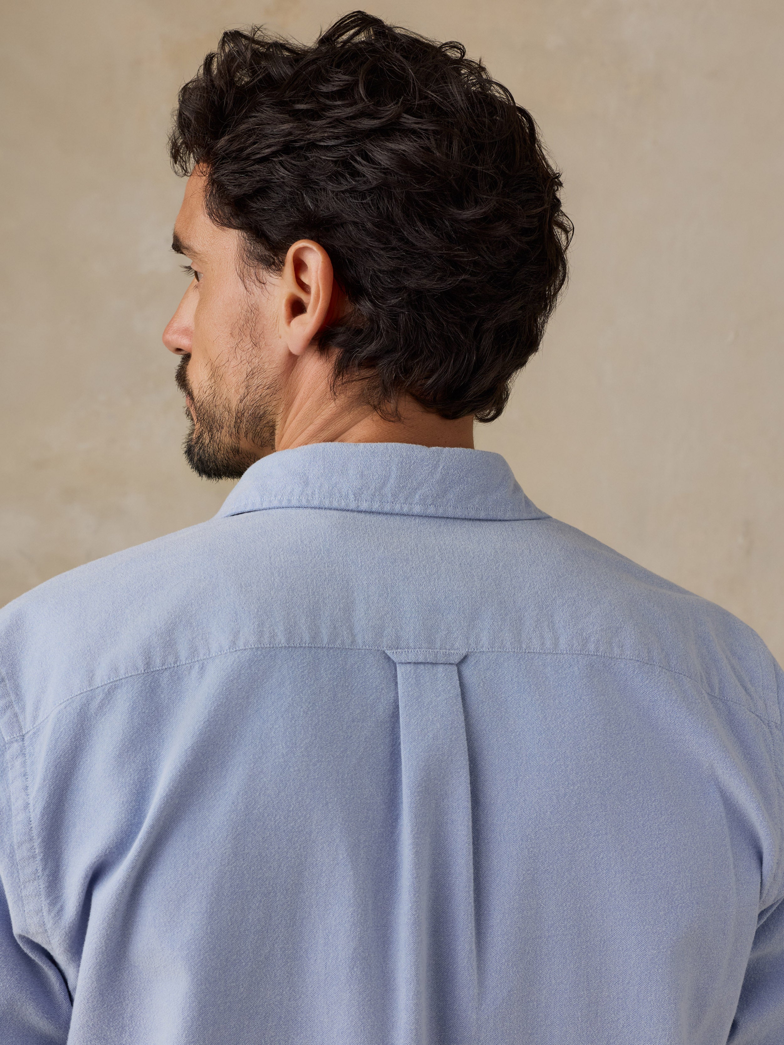 A man shown from the back wearing a light blue long-sleeve button-down shirt against a neutral beige background., image 6 of 9