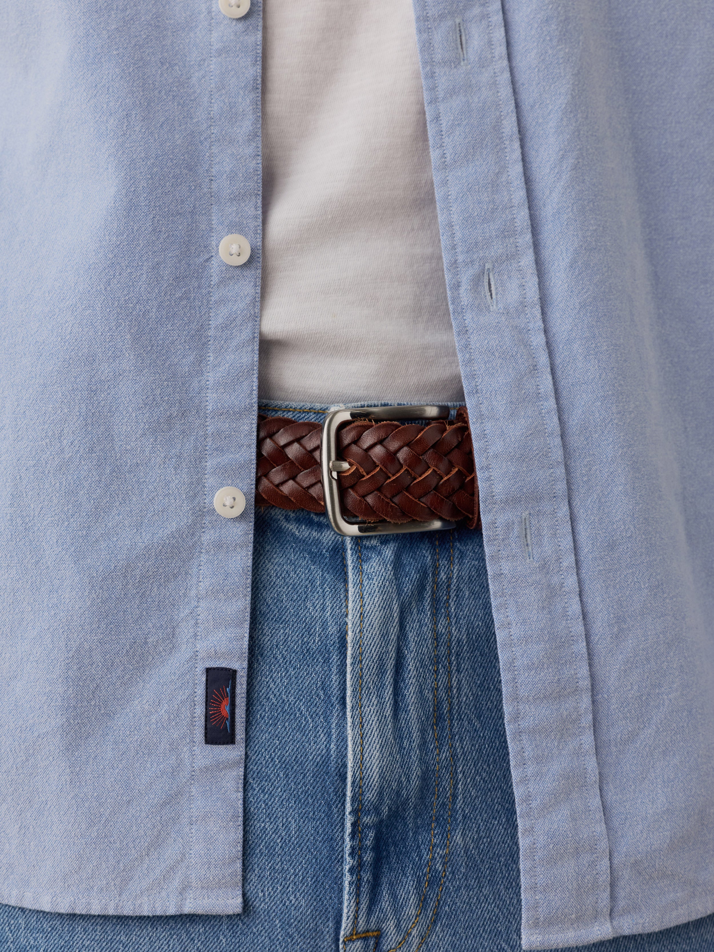 Close-up of a man wearing a light blue button-up shirt, a cream undershirt, blue jeans, and a brown braided belt with a silver buckle., image 4 of 9