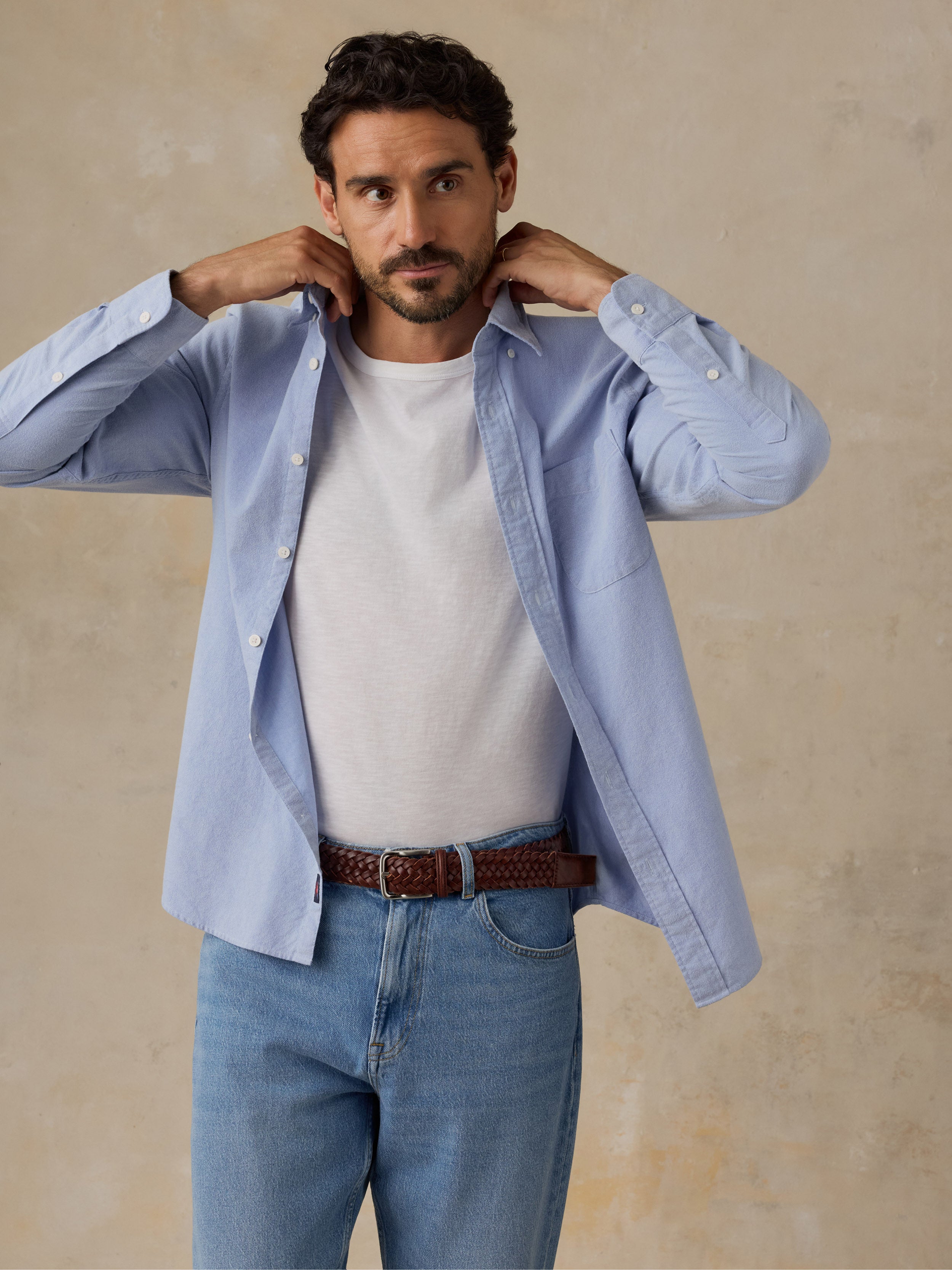 A man wearing a light blue long-sleeve button-down shirt over a white t-shirt, paired with blue jeans and a brown belt, posing against a textured beige background., image 5 of 9