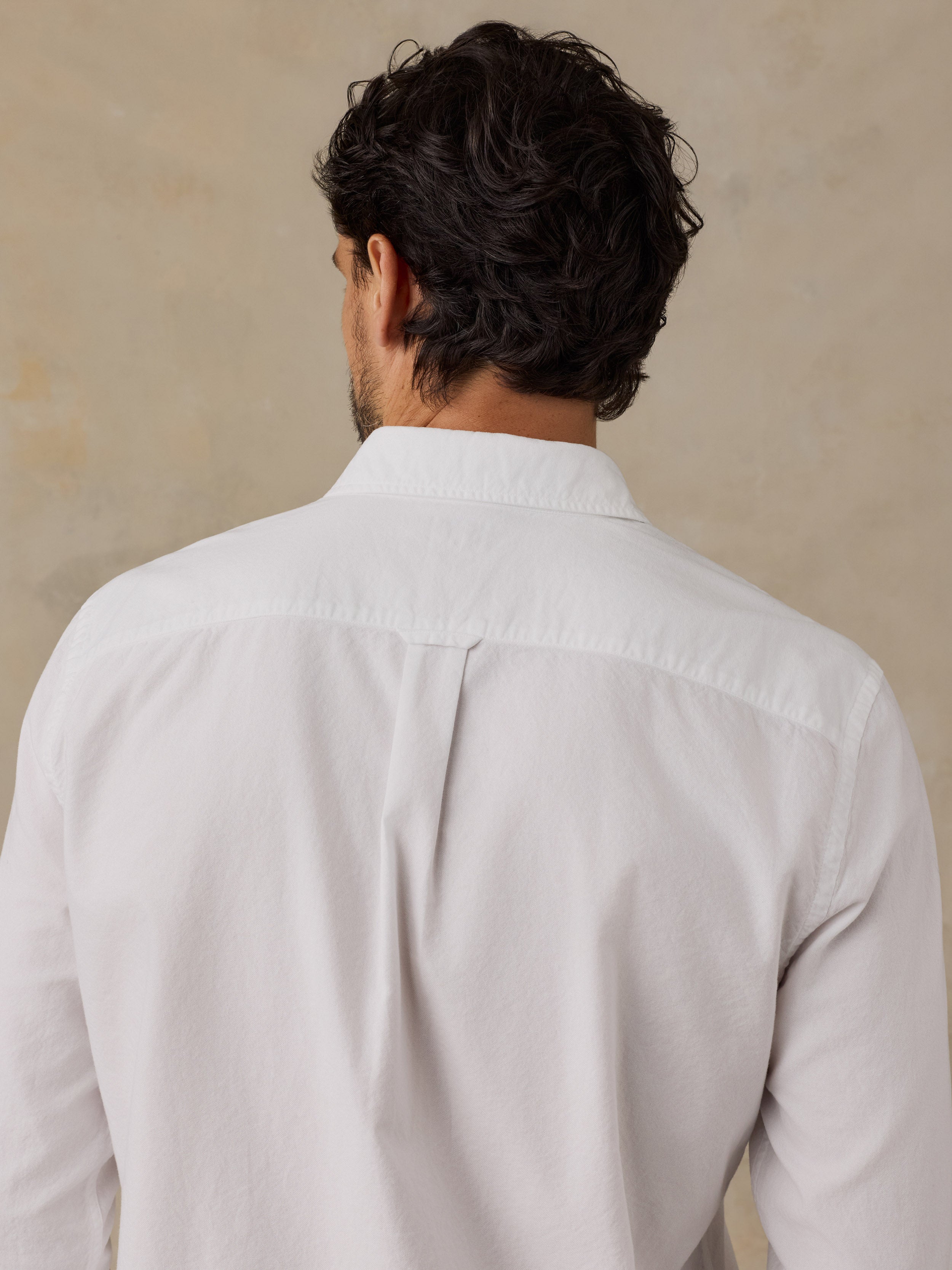 A man viewed from behind wearing a white long-sleeve button-down shirt, showing the back collar and shoulder seam details against a neutral beige background.