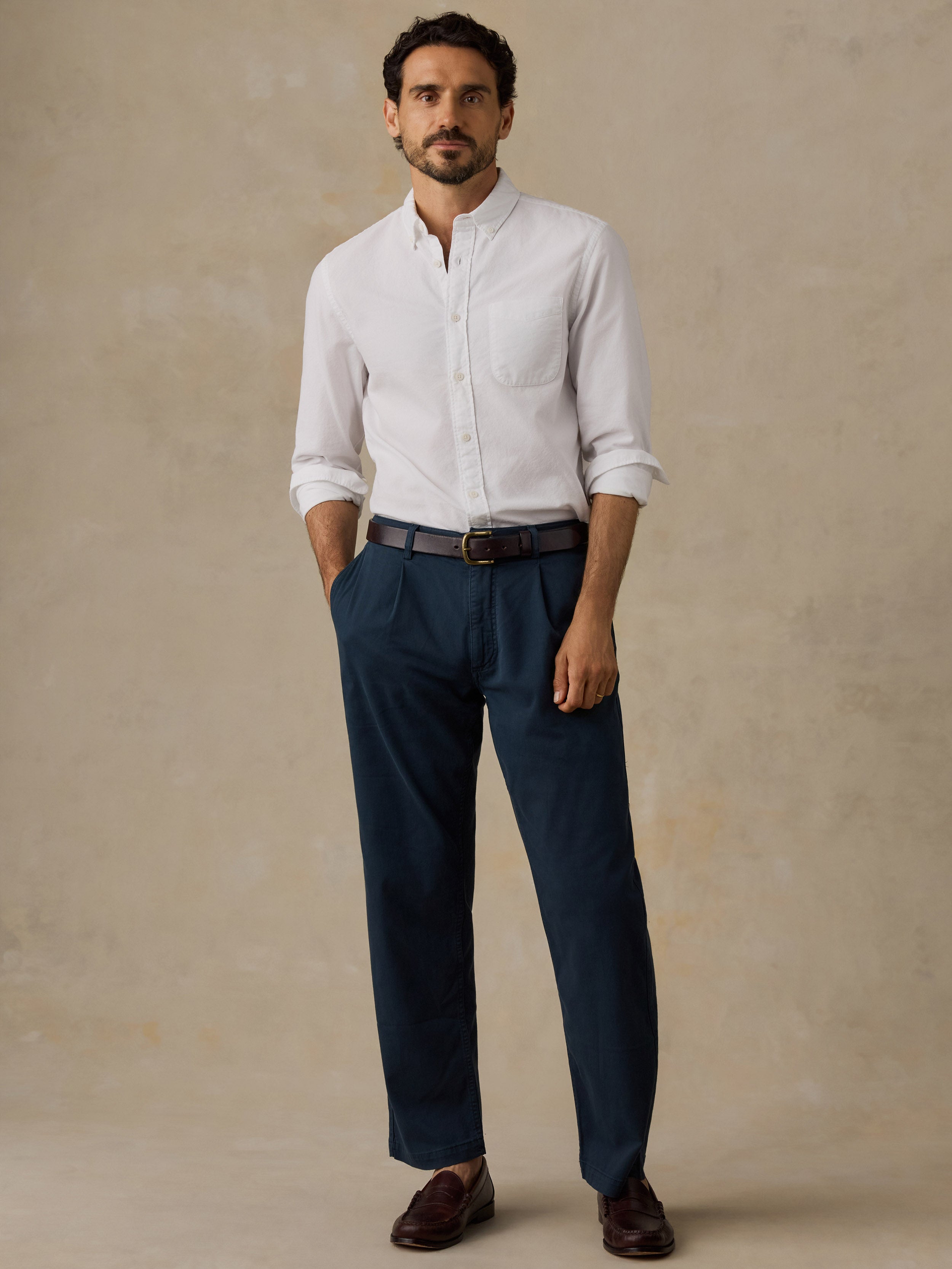 A man wearing a white long-sleeve button-down shirt, navy blue trousers, a dark brown belt, and dark brown loafers, standing against a textured beige studio backdrop.