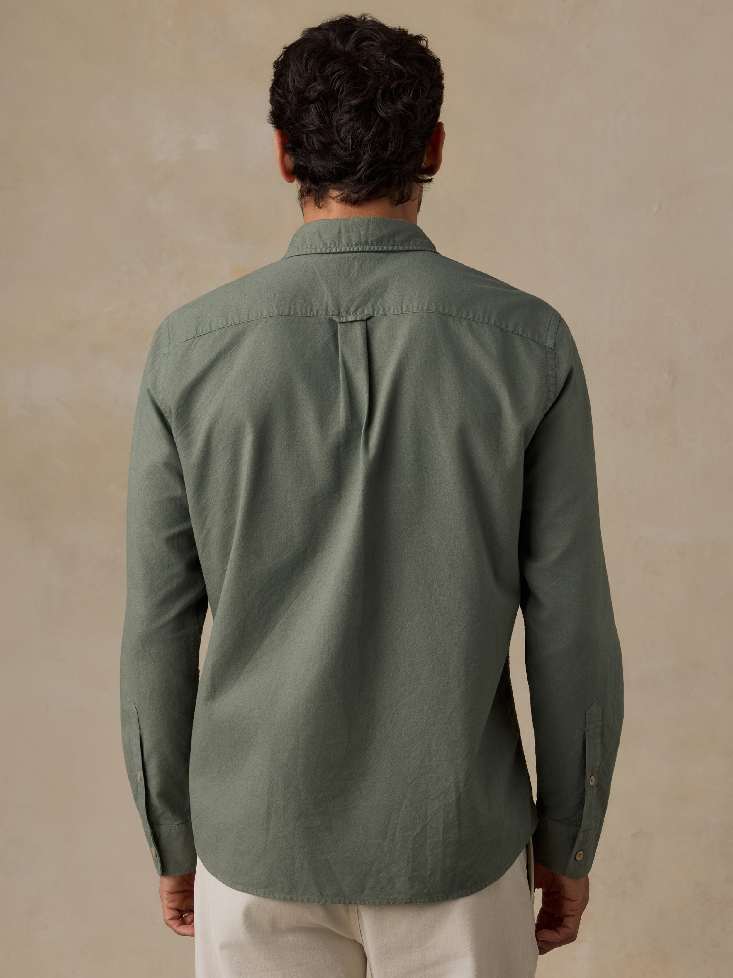 A man viewed from behind wearing a muted sage green long-sleeve button-up shirt and off-white trousers, standing against a textured beige wall.