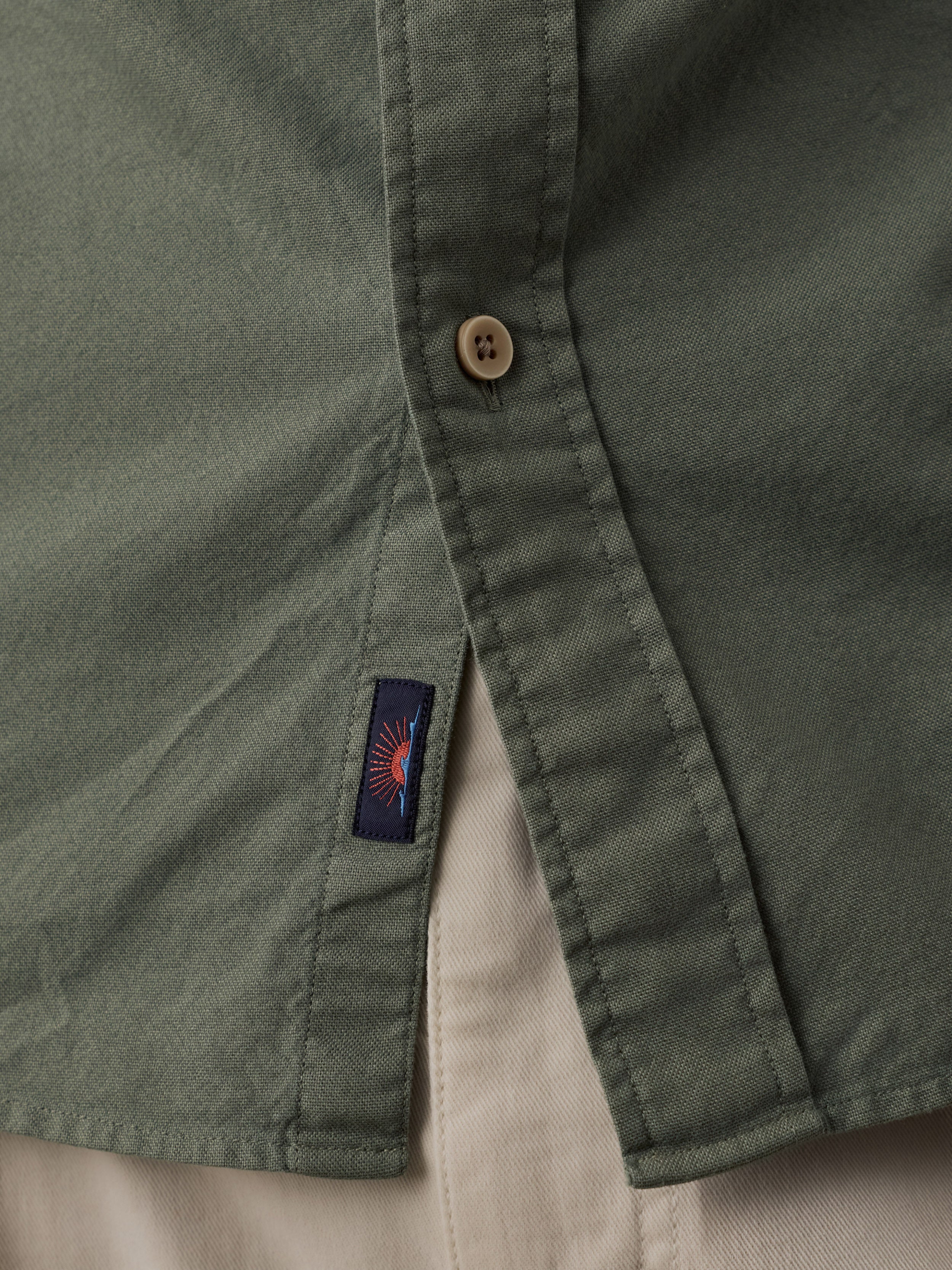 Close-up of an olive green button-up shirt showing the button closure and a small navy Faherty brand label with a sun and waves logo.