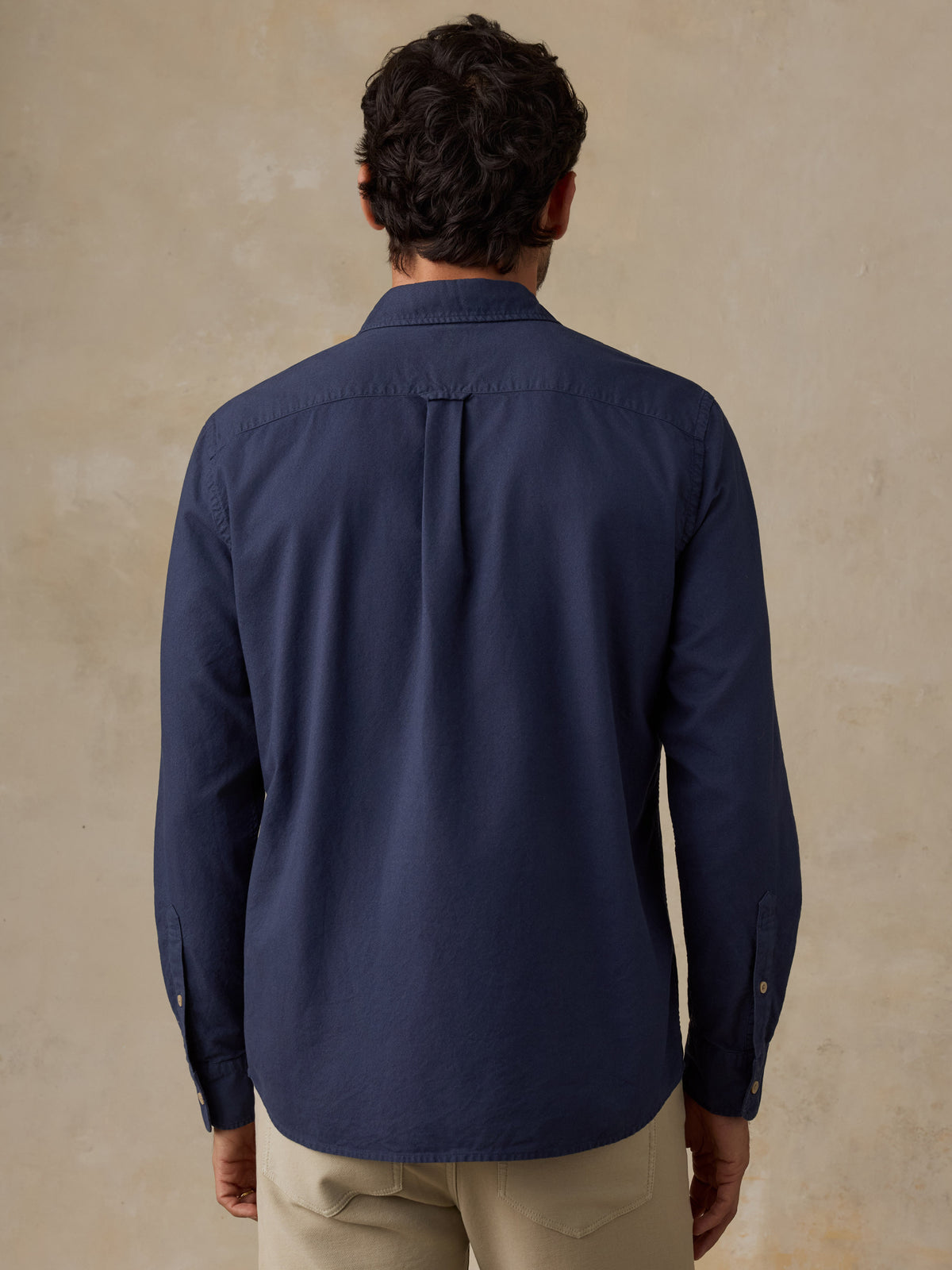 A man viewed from behind wearing a navy blue long-sleeve button-down shirt and beige trousers, standing against a textured beige wall., image 3 of 9
