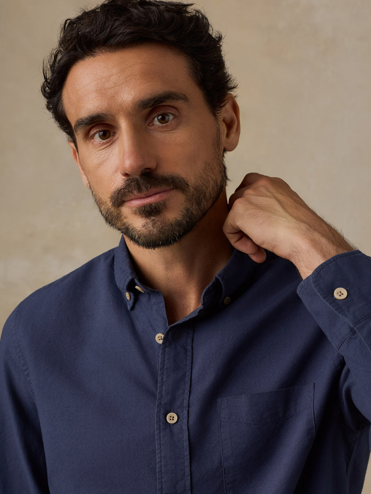 A close-up portrait of a man wearing a navy blue long-sleeve button-down shirt against a textured beige background., image 6 of 9
